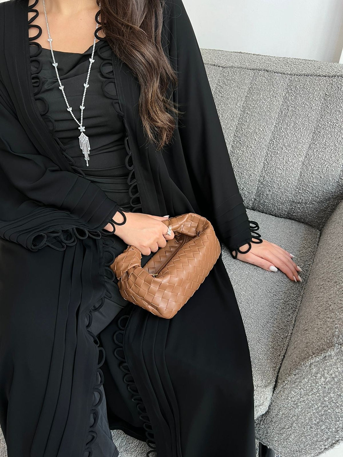 Person wearing a black outfit with ruffled details, holding a brown handbag on a gray couch.