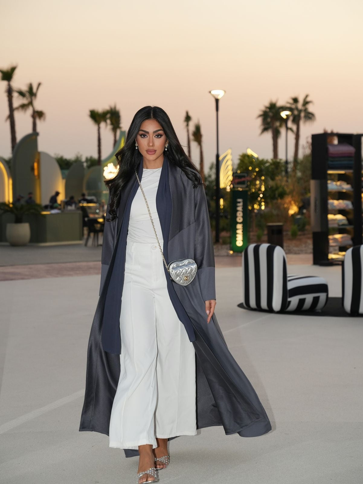 Woman in a long dark coat over a white outfit walking outdoors with palm trees and modern architecture in the background.