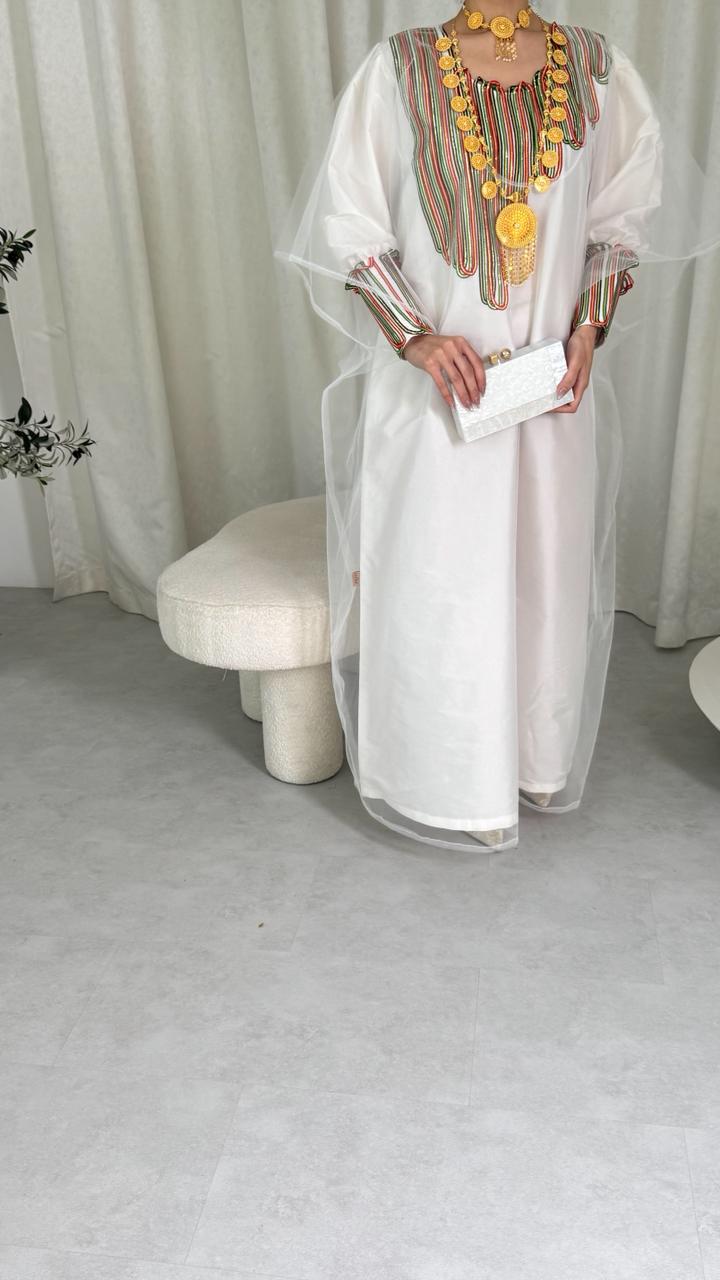 Person wearing a white dress with colorful embroidery and accessories, standing against a plain background.