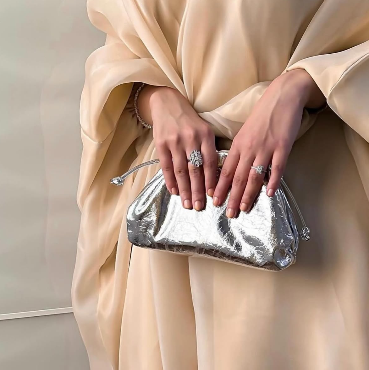 Person holding a silver clutch bag in a beige outfit on a neutral background