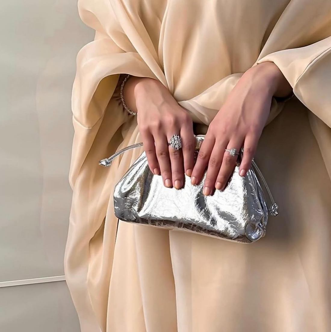 Person holding a silver clutch bag in a beige outfit on a neutral background