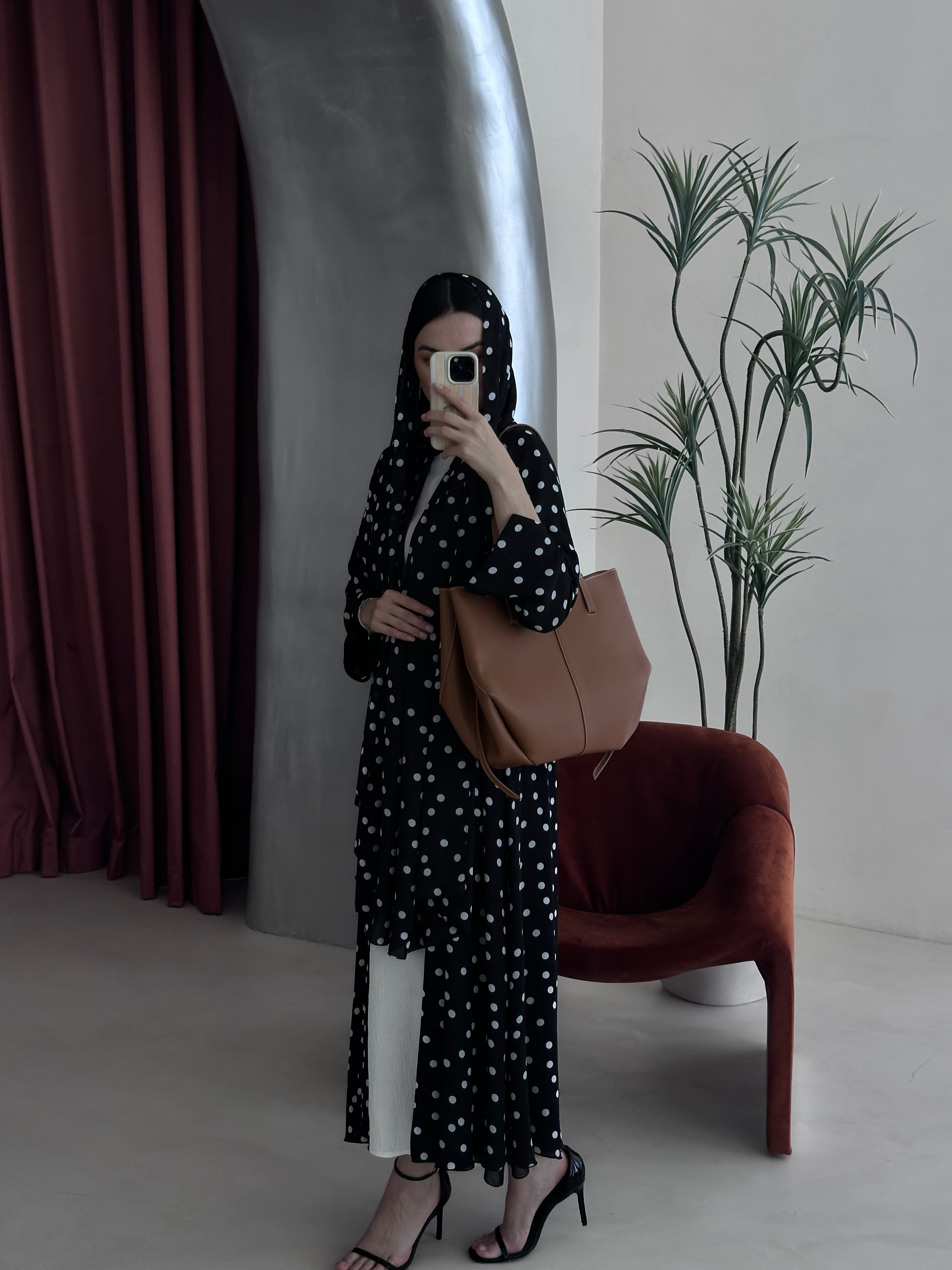 Woman in a polka dot dress holding a brown bag in a room with a plant and chair.