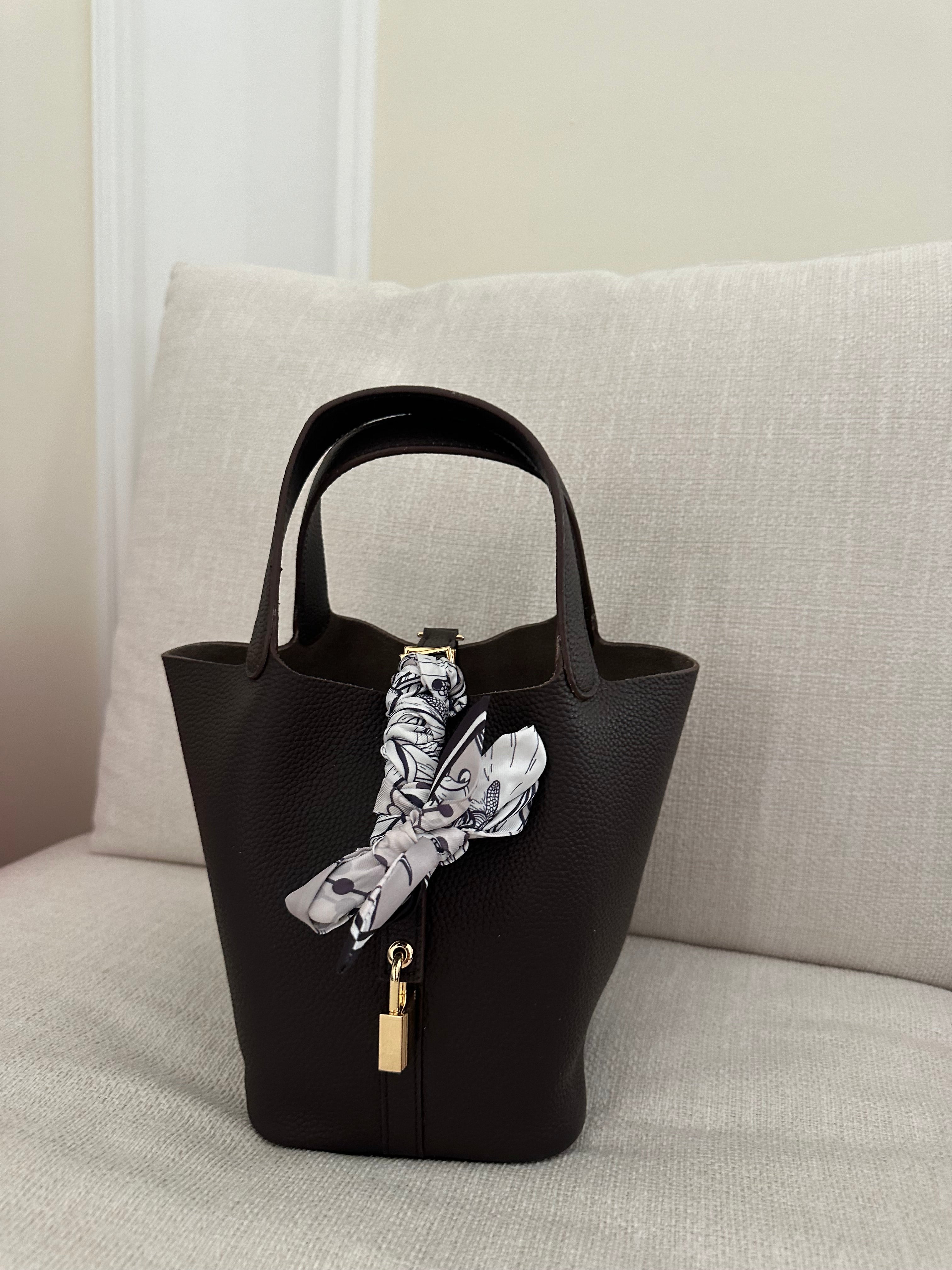 A black handbag with a burgundy detail, displayed on a white sofa. It features a metallic clasp, a hanging lock, and a patterned scarf attached to it.