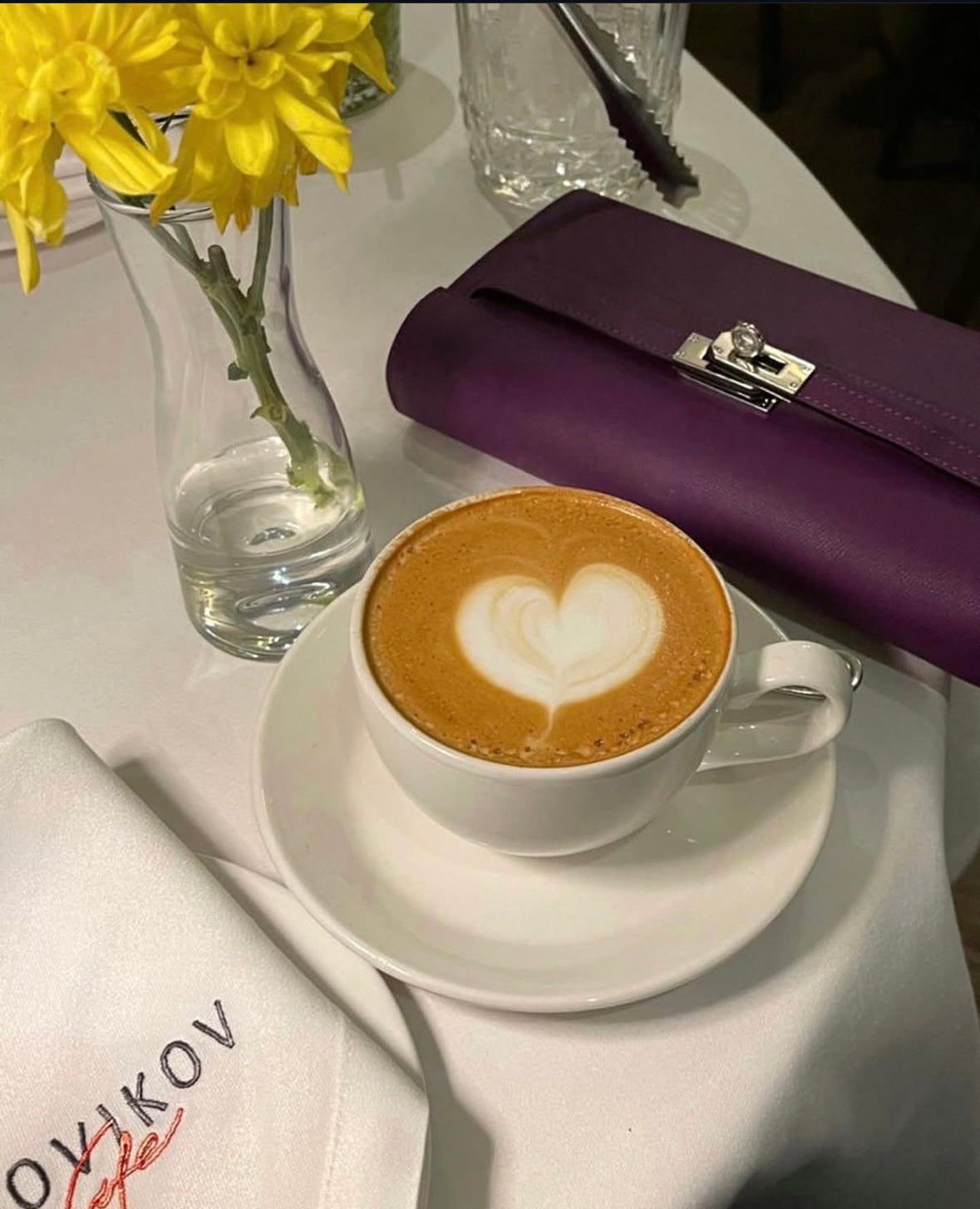 Cappuccino with heart foam art on a white saucer, purple clutch, and yellow flowers in the background.
