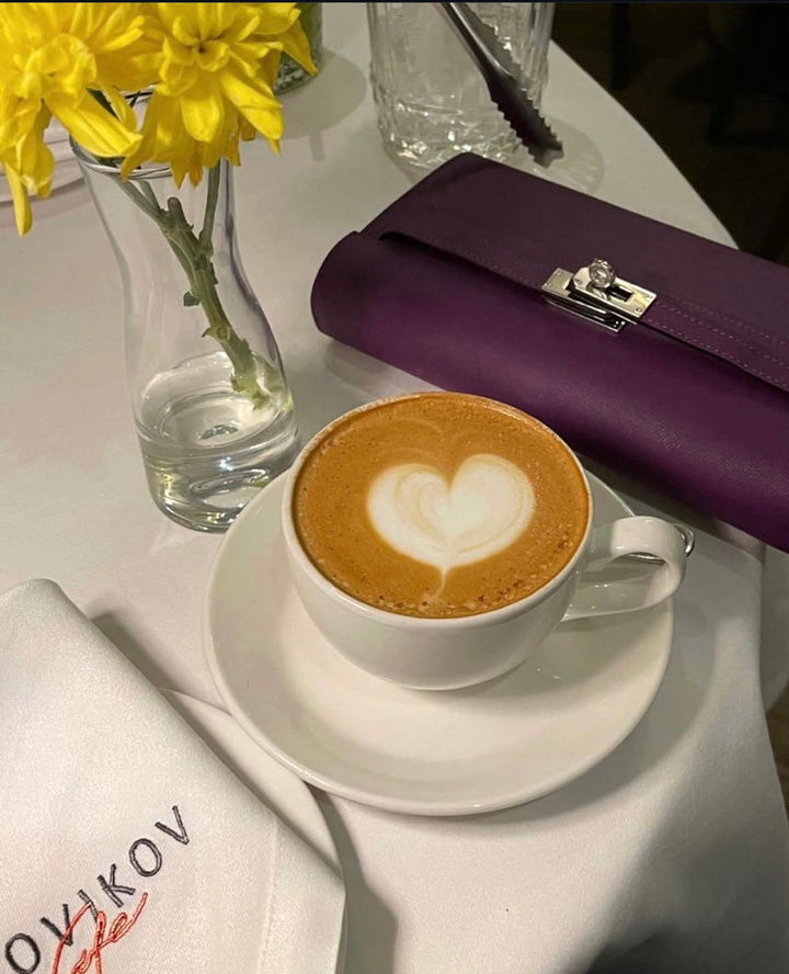Cappuccino with heart foam art on a white saucer, purple clutch, and yellow flowers in the background.