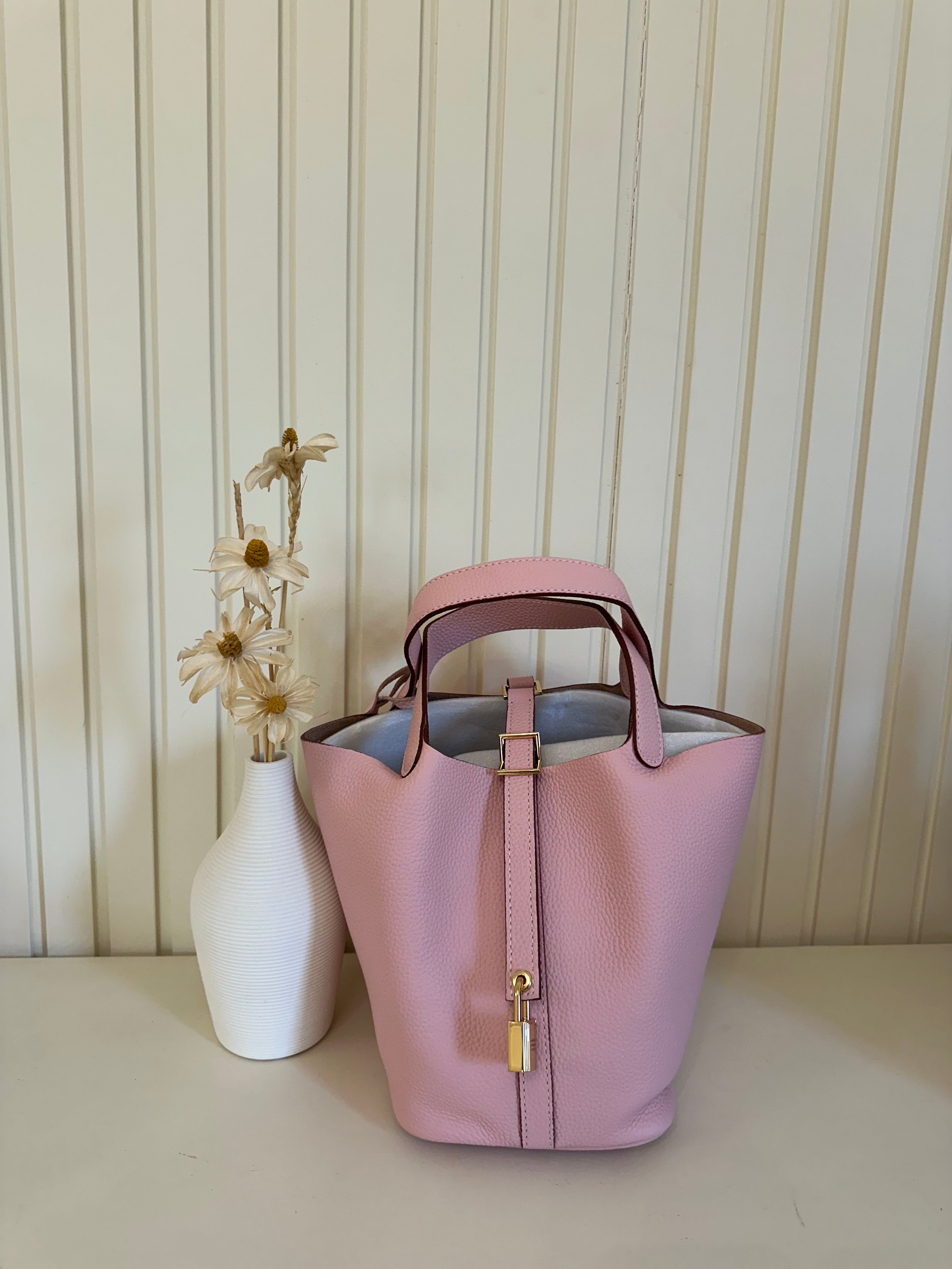 A pink handbag with a gold finish padlock, displayed alongside a white vase with dried flowers.