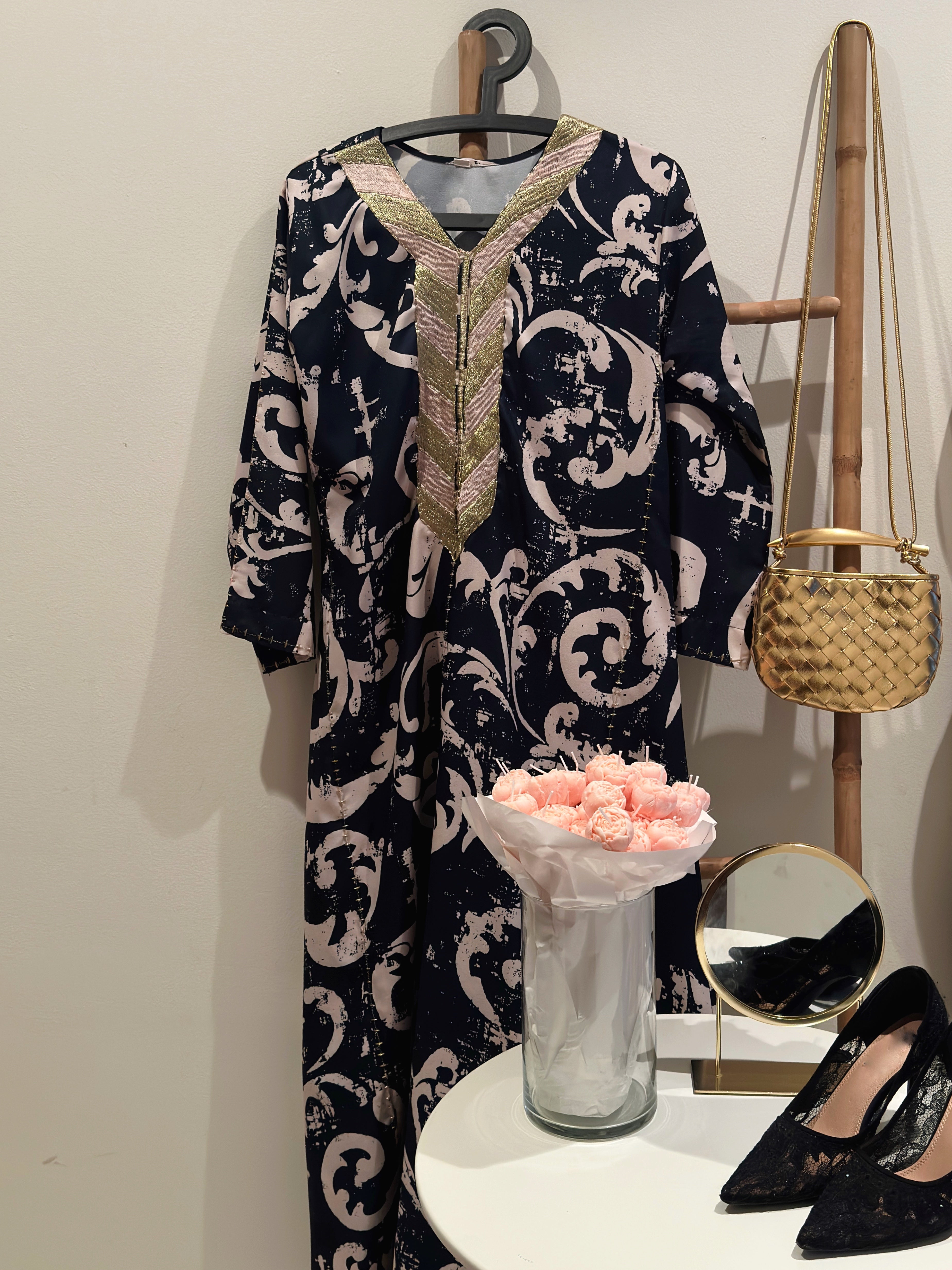 A navy and gold patterned handbag displayed on a mannequin, alongside a vase with pink flowers and black high-heeled shoes.
