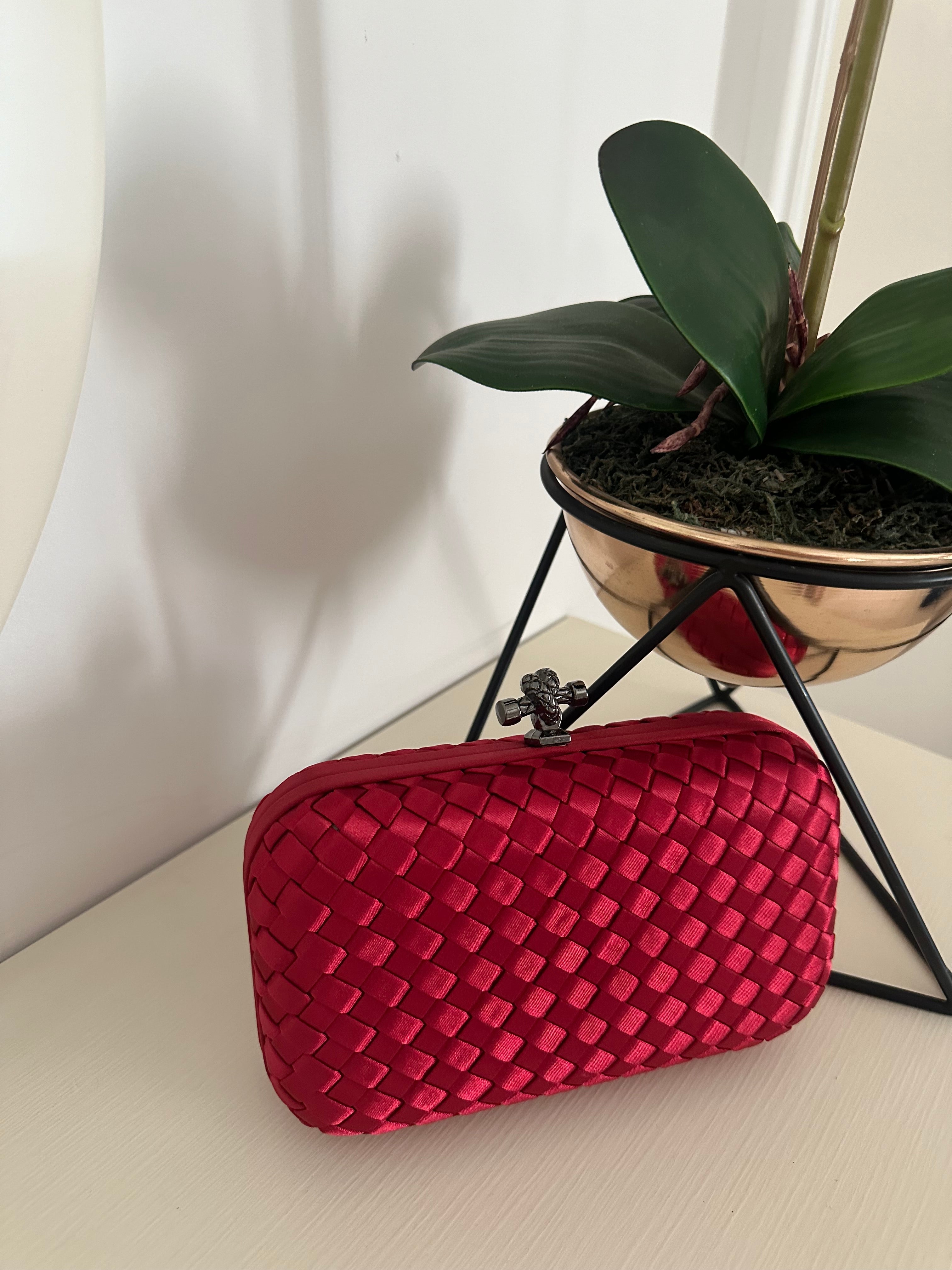A red clutch purse with a geometric pattern, placed on a metal stand, with a plant in the background.