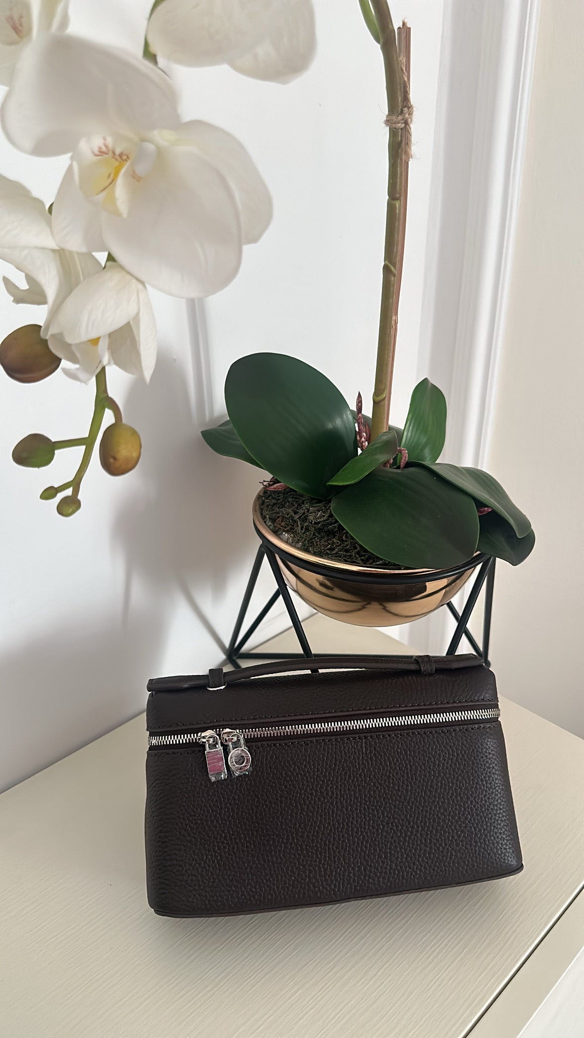 Black handbag with silver zippers next to a potted orchid on a light surface.