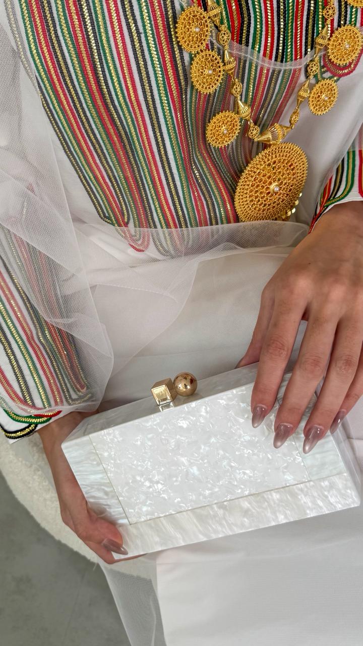 Person holding a white clutch with gold accents, wearing a colorful embroidered top.