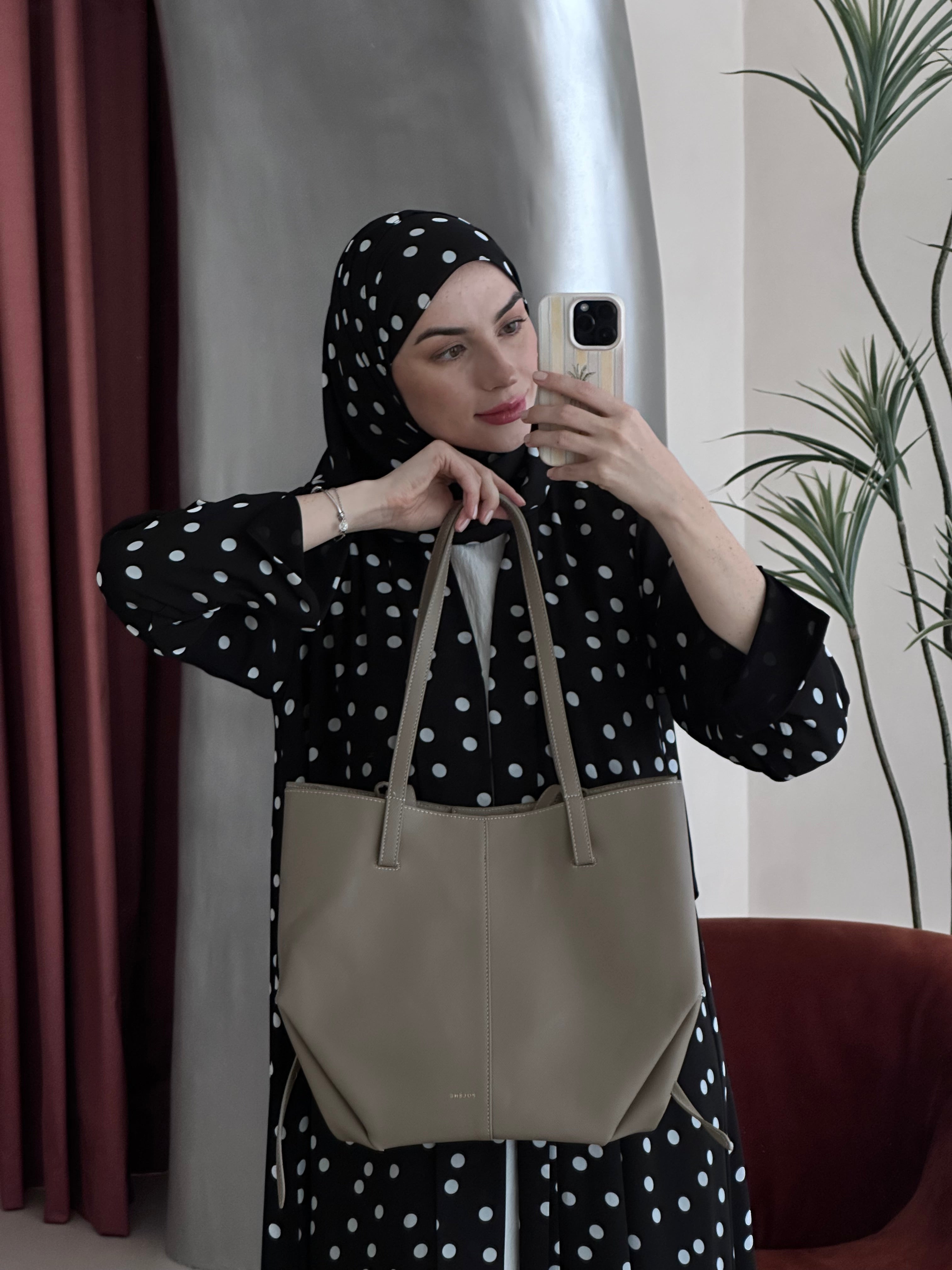 Woman taking a mirror selfie wearing a black polka dot hijab and holding a beige tote bag.