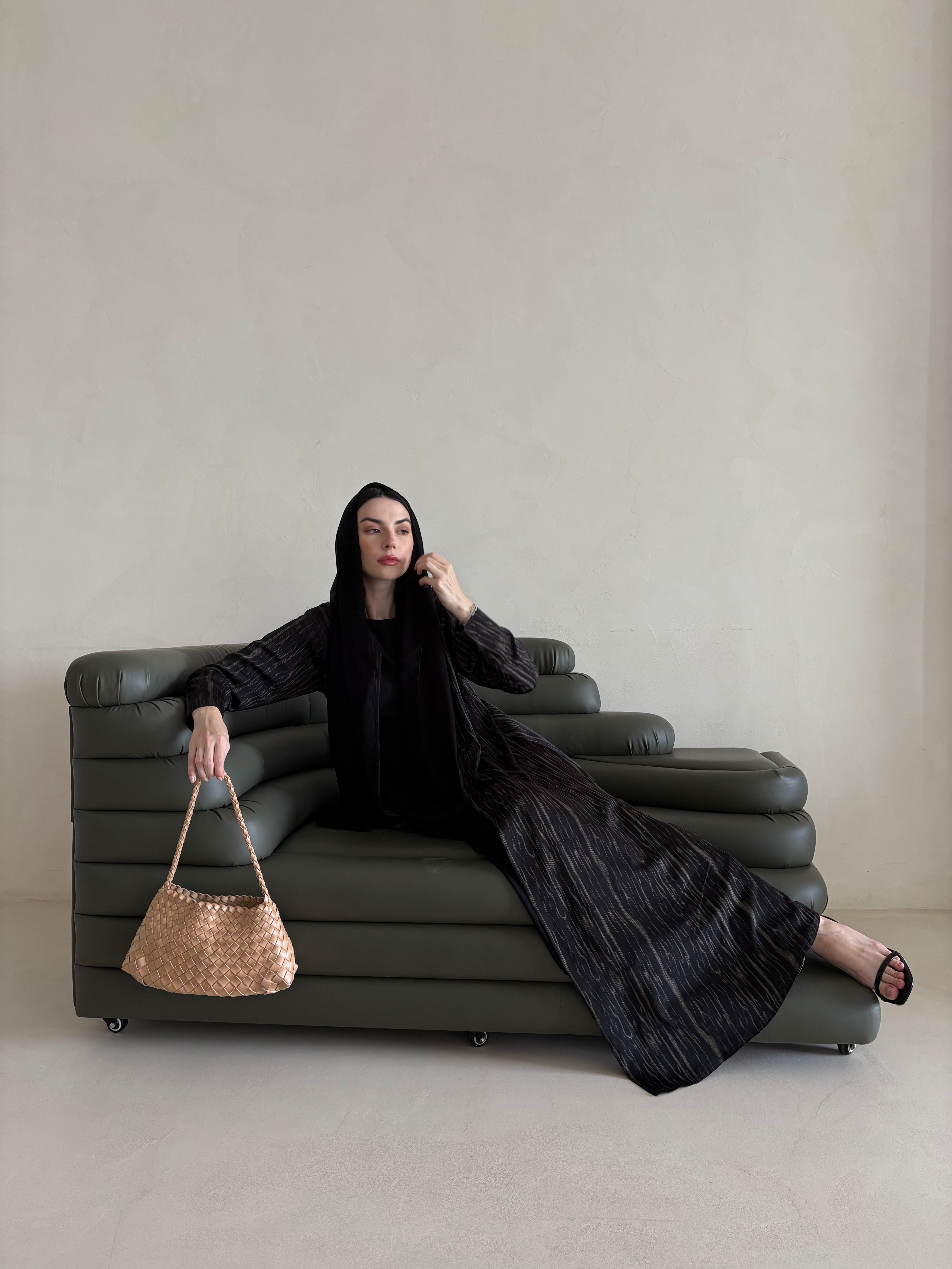 Woman in a black abaya sitting on a modern sofa holding a woven handbag.