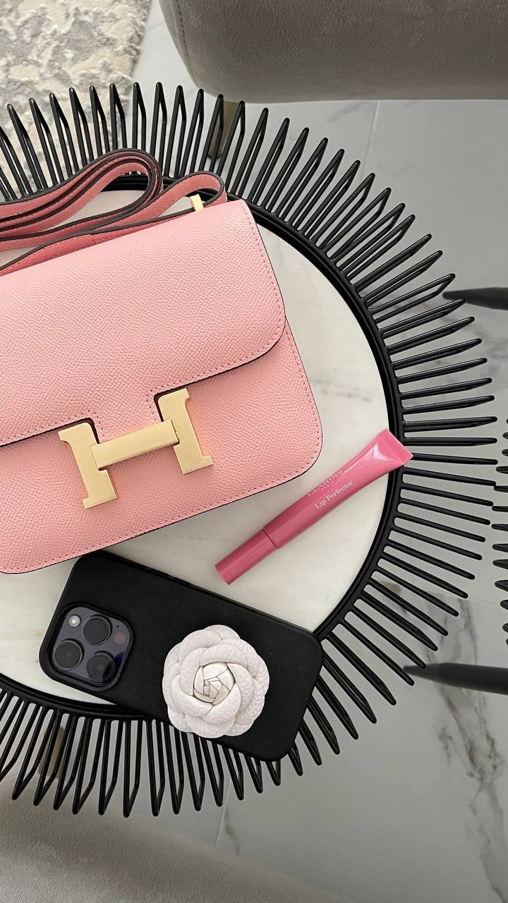 Pink handbag with gold 'H' logo, black comb, pink marker, and black phone case on a marble surface.