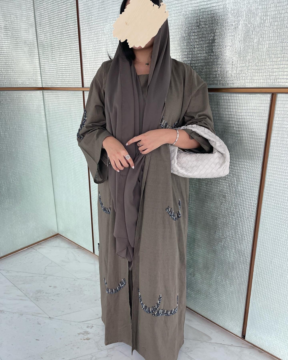 Person wearing a long brown dress with embroidery in front of a textured wall.