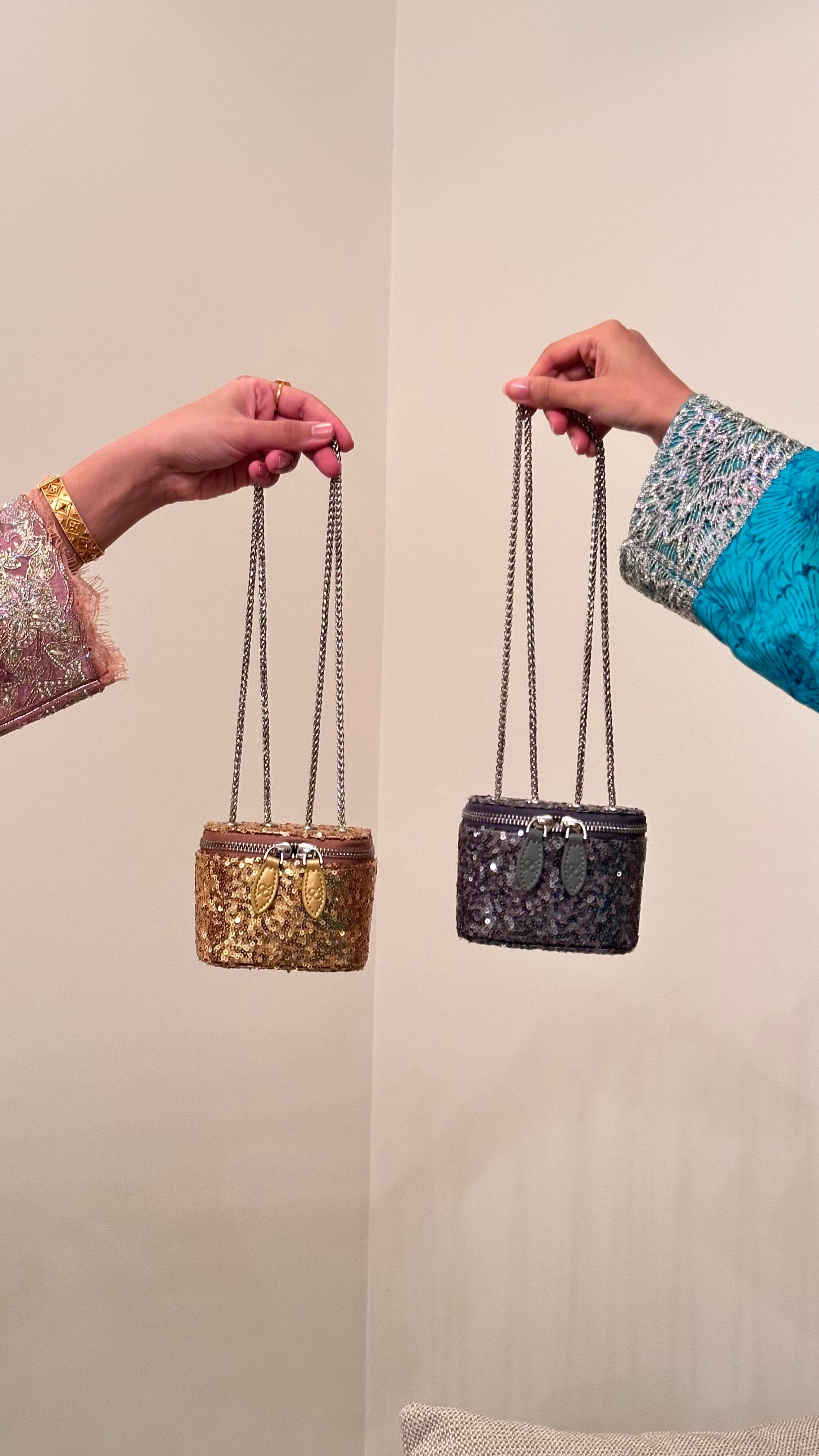 Two hands holding a gold and a silver handbag against a neutral background