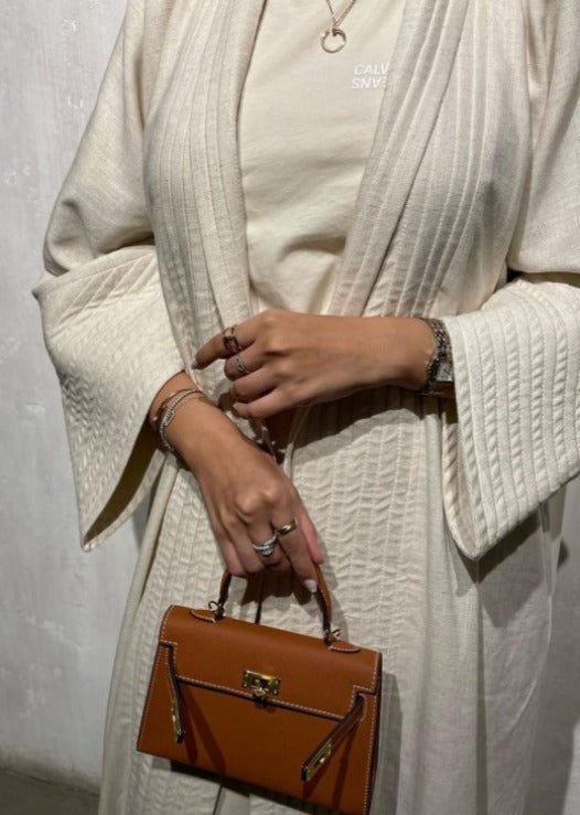 A person wearing a white coat holding a brown leather handbag with a strap.