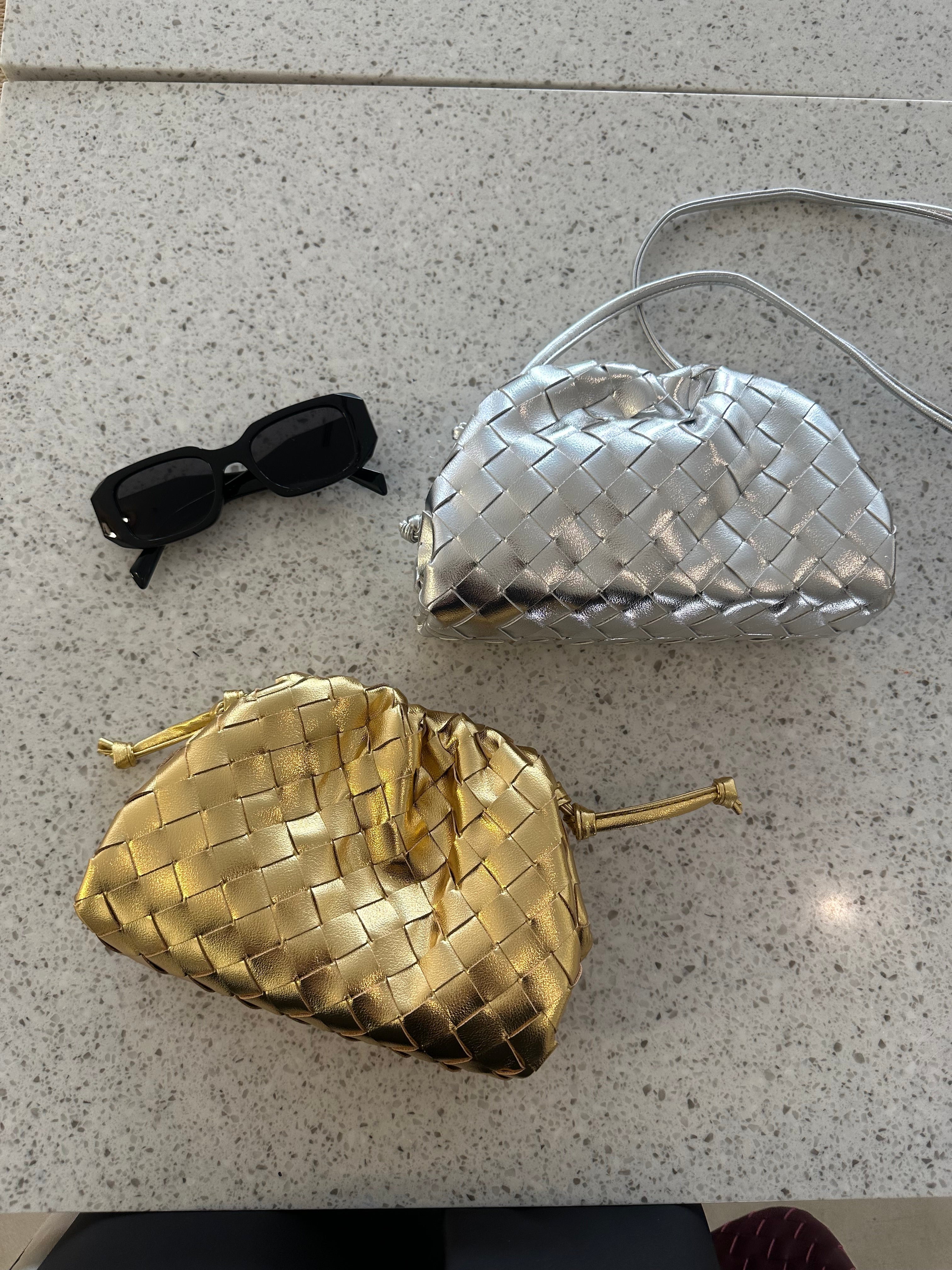 Two interwoven handbags in gold and silver colors, alongside a pair of sunglasses, displayed on a surface.