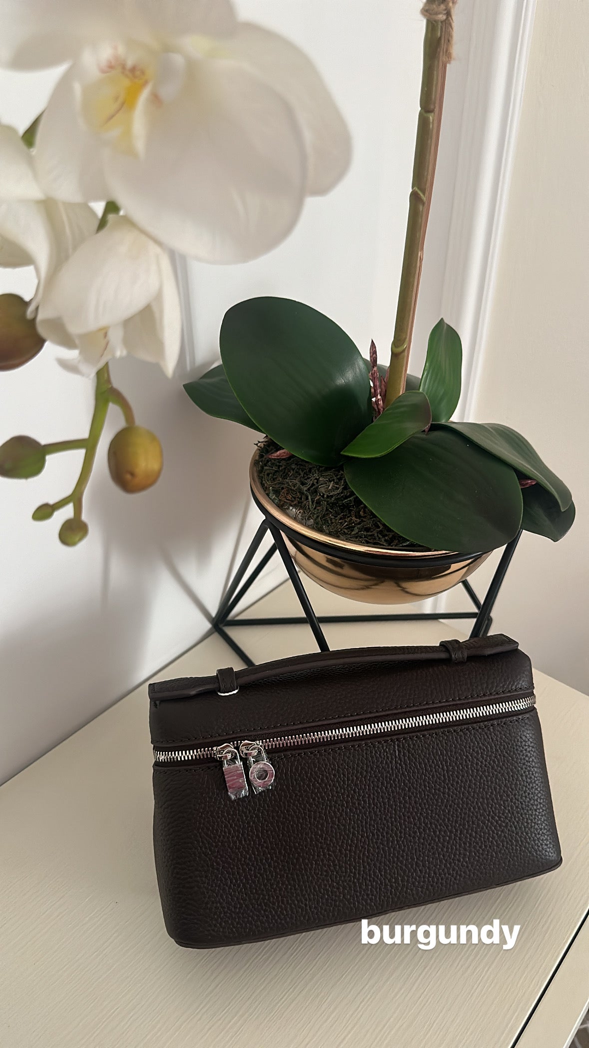 Black handbag with silver zipper on a white surface with a plant and white flowers in the background.