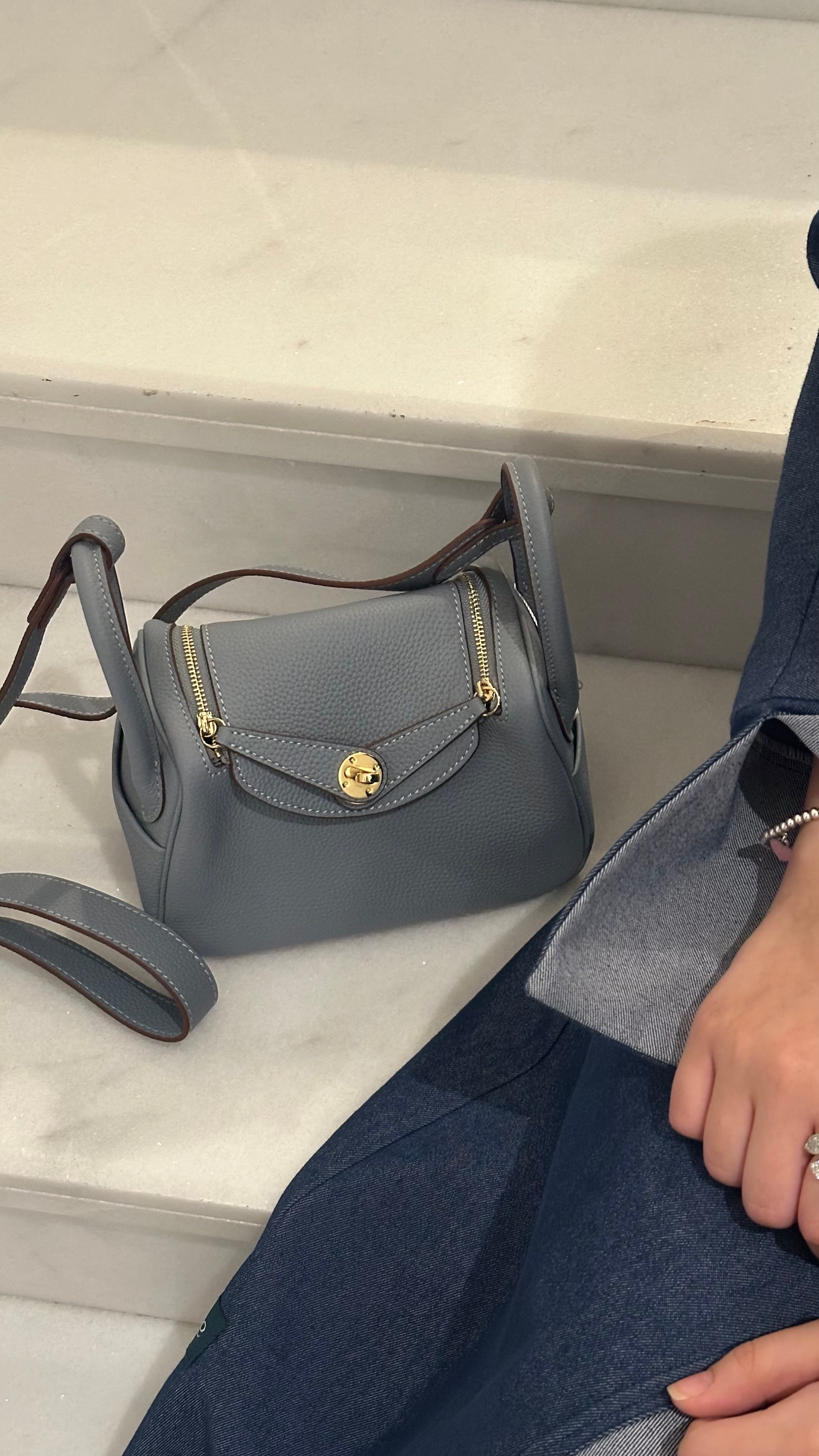 A light blue leather clutch bag with a luxury design, placed on a marble surface with a person's hand partially visible.