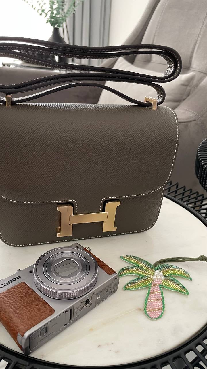 Brown handbag with gold clasp on a marble surface next to a camera and palm tree decoration.