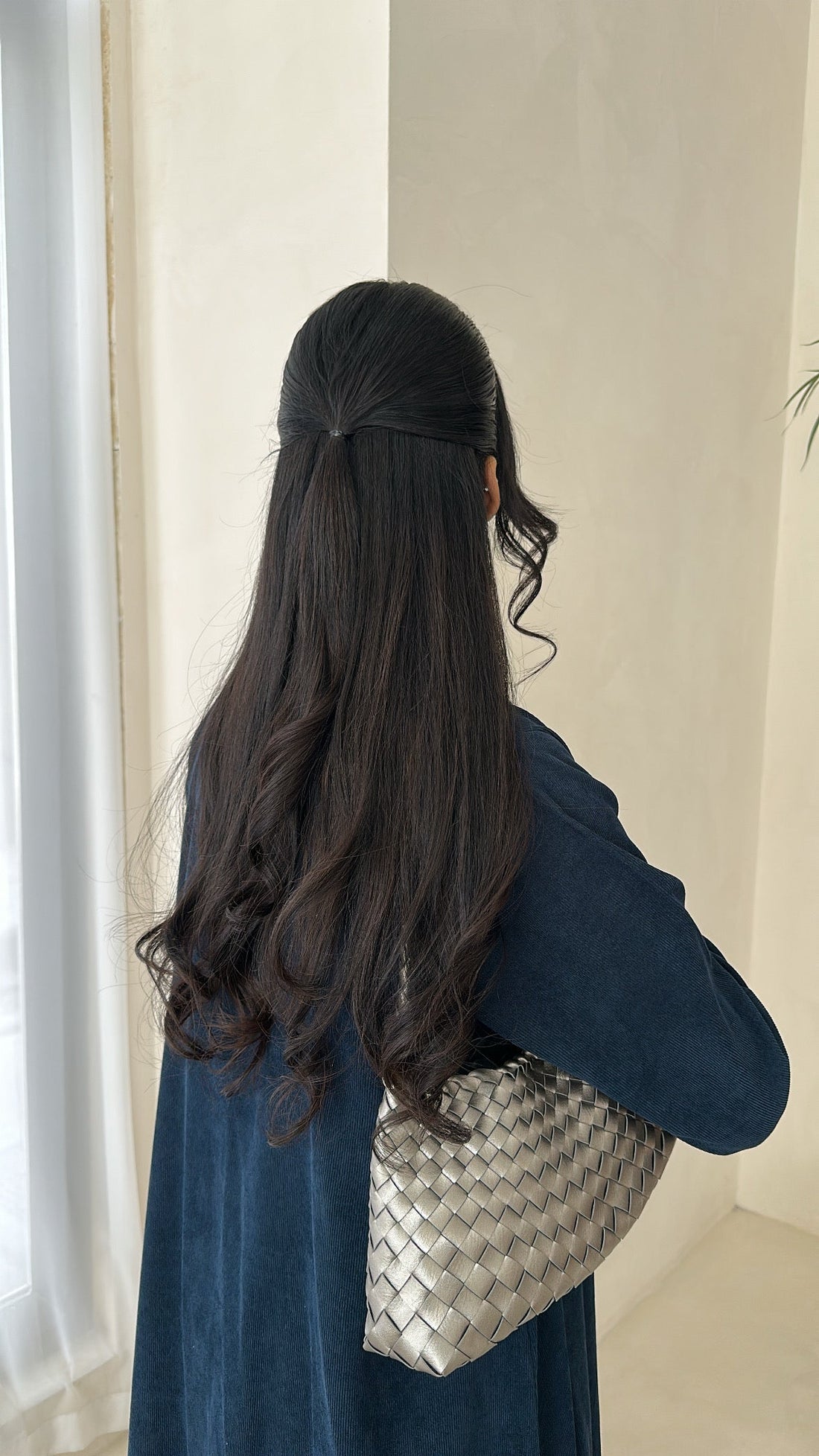 Woman with long dark hair wearing a blue dress holding a silver handbag indoors.