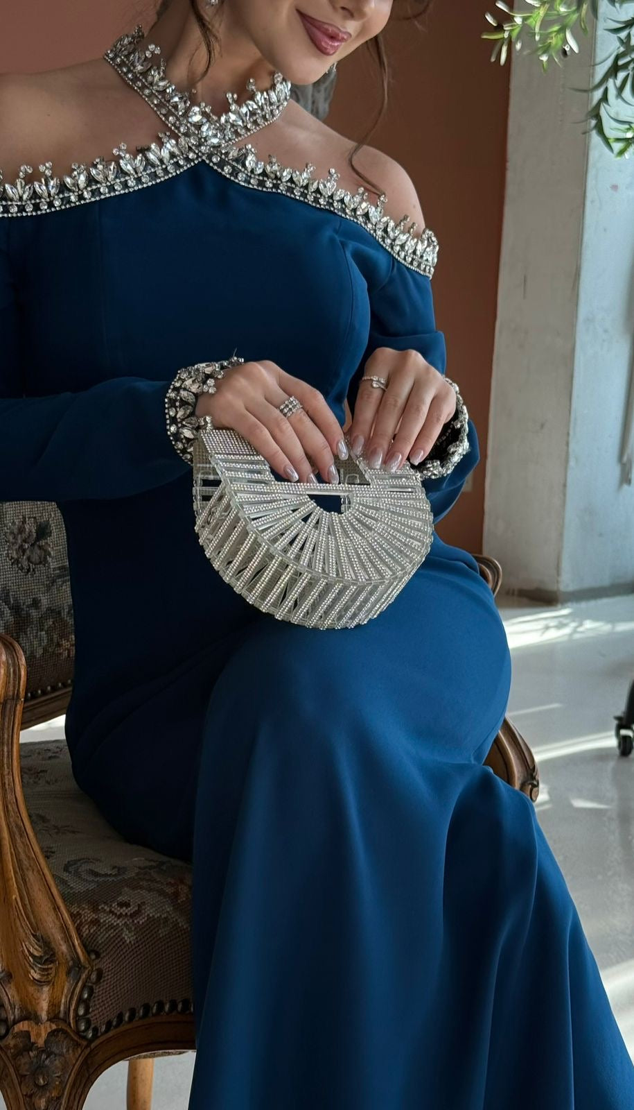Woman in a blue evening gown with a beaded clutch sitting on a chair.