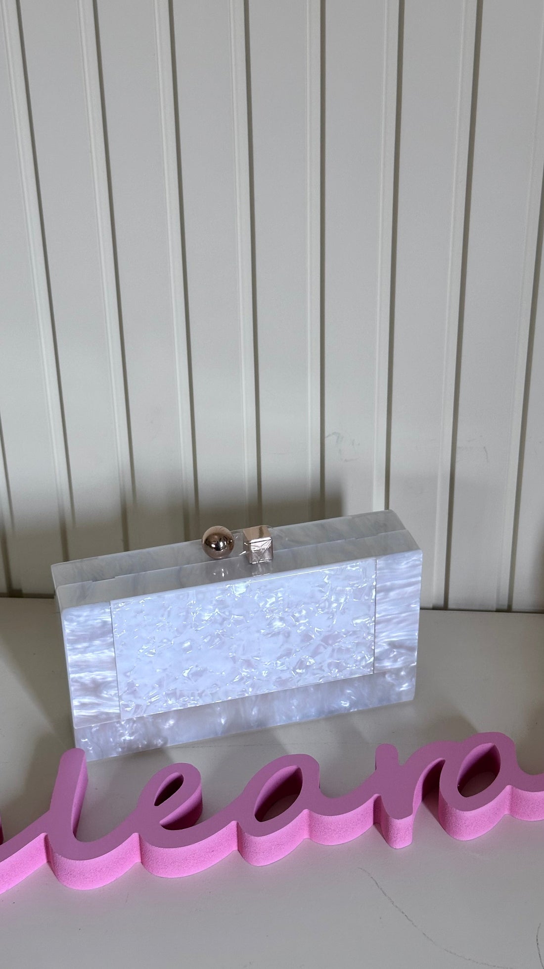 Clear clutch bag with silver accents on a white surface with pink letters spelling 'leona'.