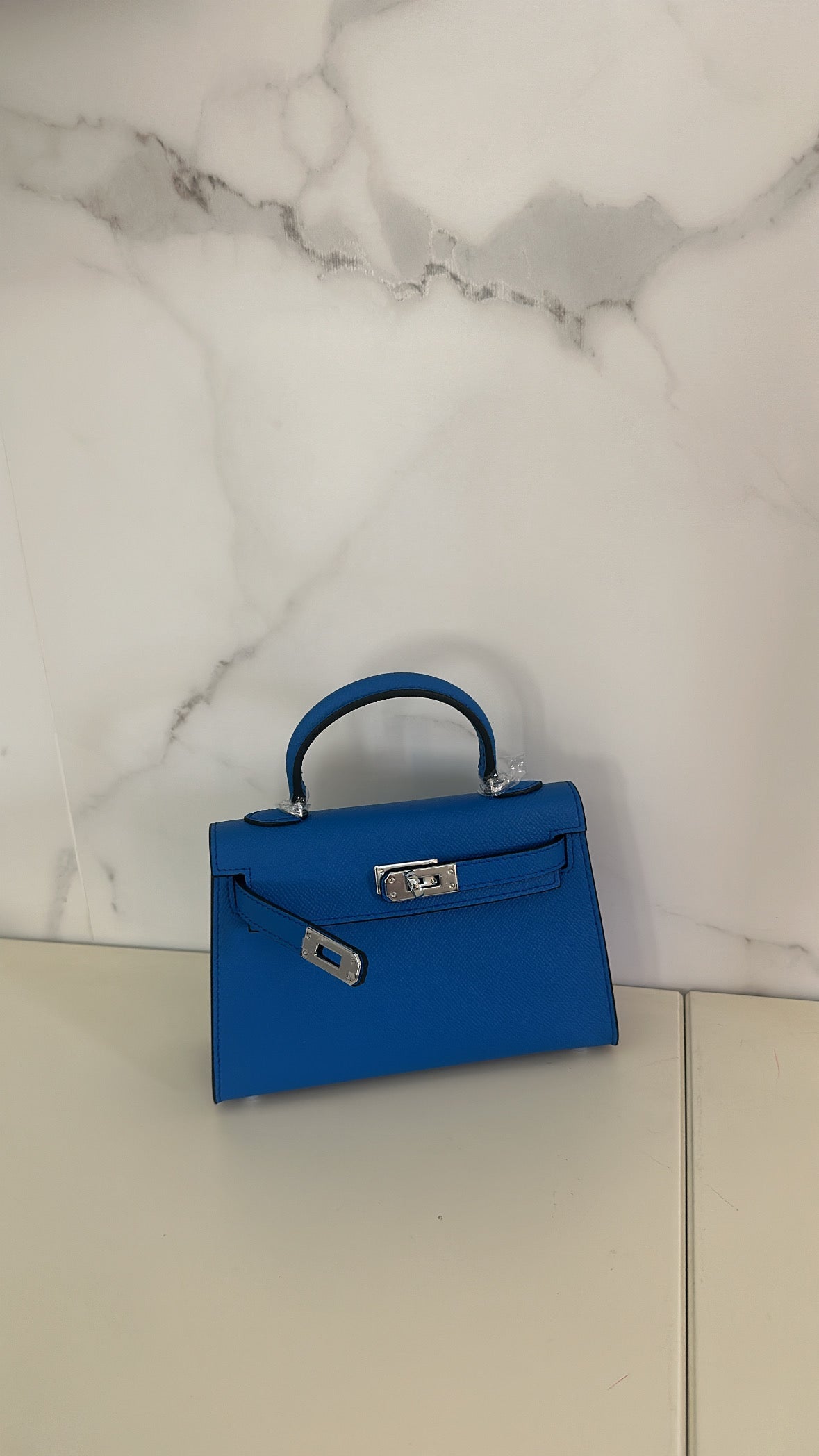 A dark blue handbag placed against a marble background, with a compact design suitable for carrying a smartphone.