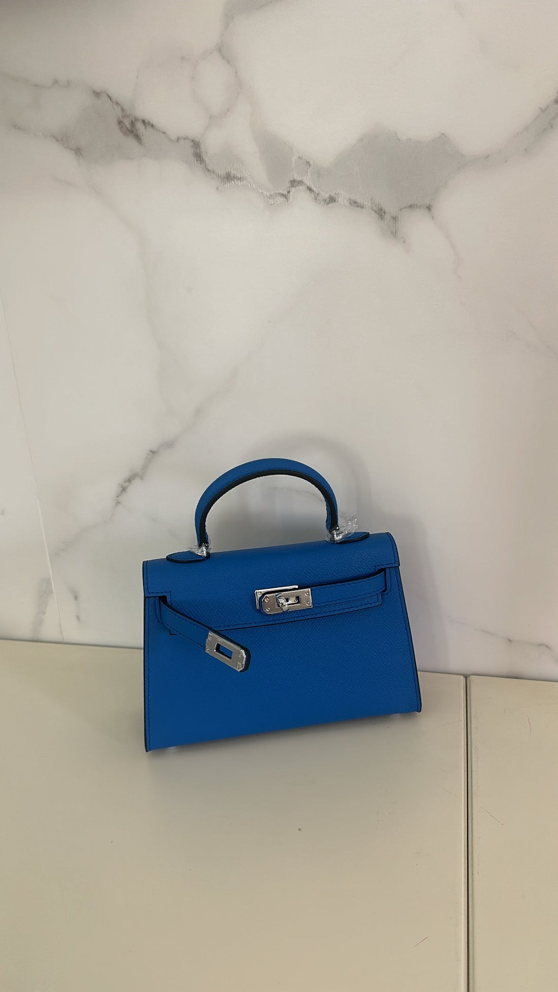 A dark blue handbag placed against a marble background, with a compact design suitable for carrying a smartphone.