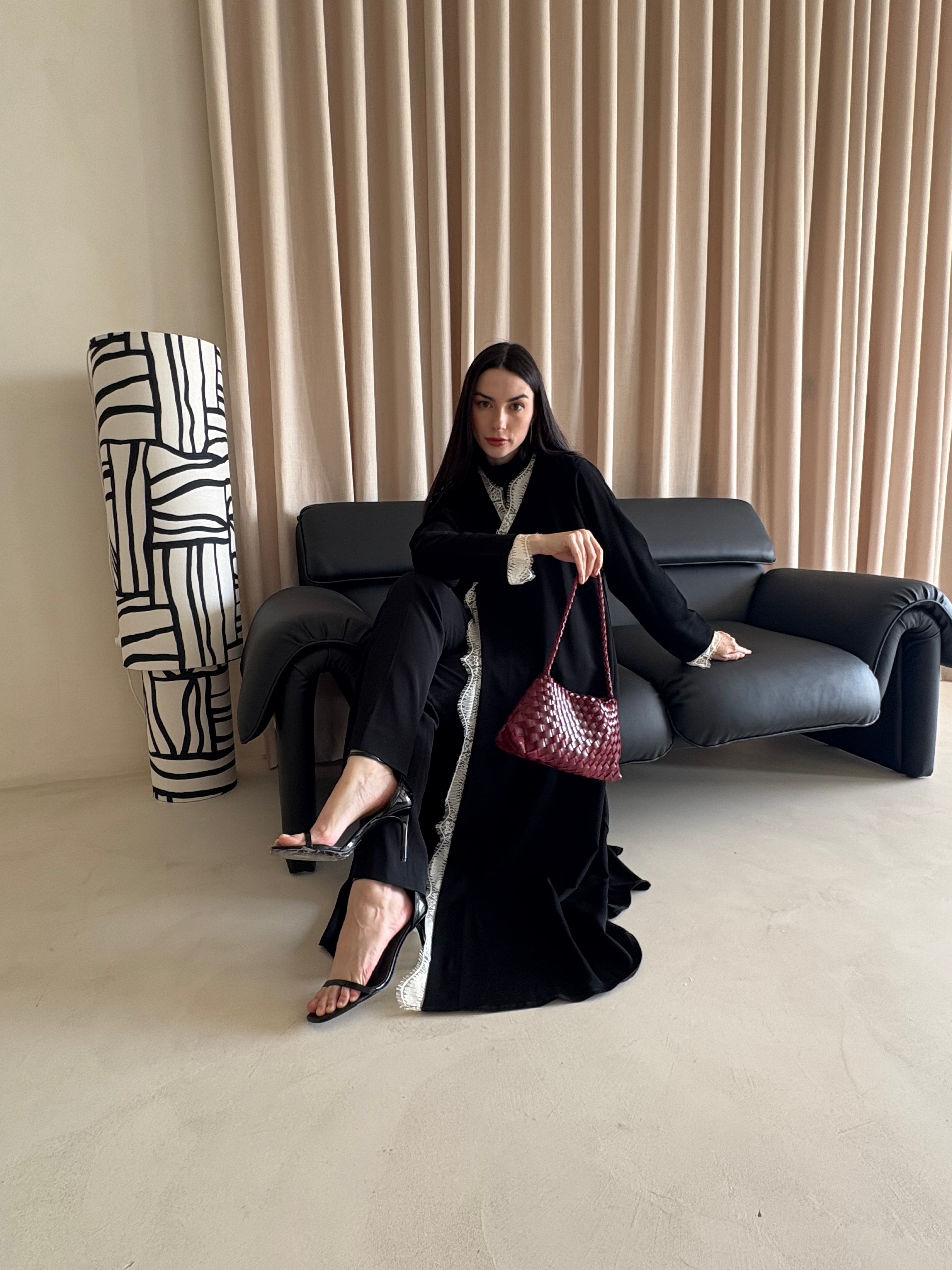 Woman in black abaya holding a red handbag in a room with a black sofa and abstract wall art.