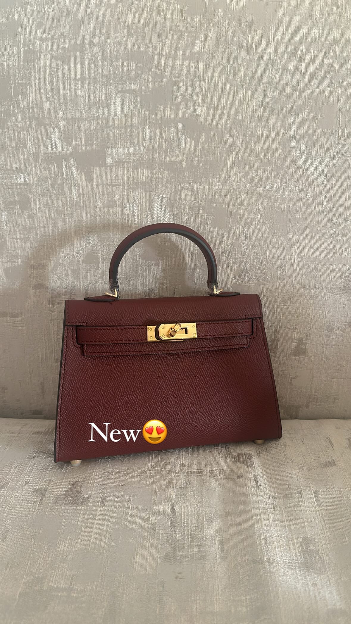 A maroon-colored handbag with a gold-tone handle and a small gold brand tag on the side, placed on a textured surface.