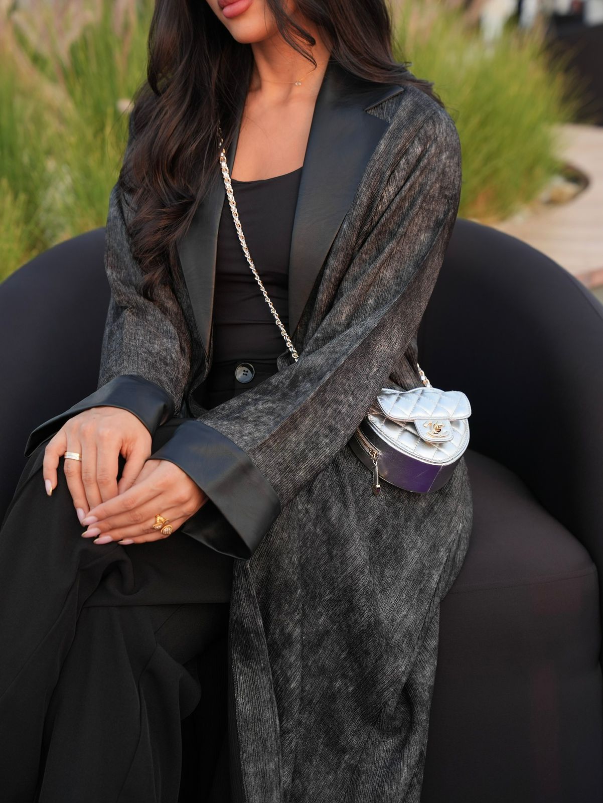 Woman wearing a gray coat over a black outfit, sitting outdoors.