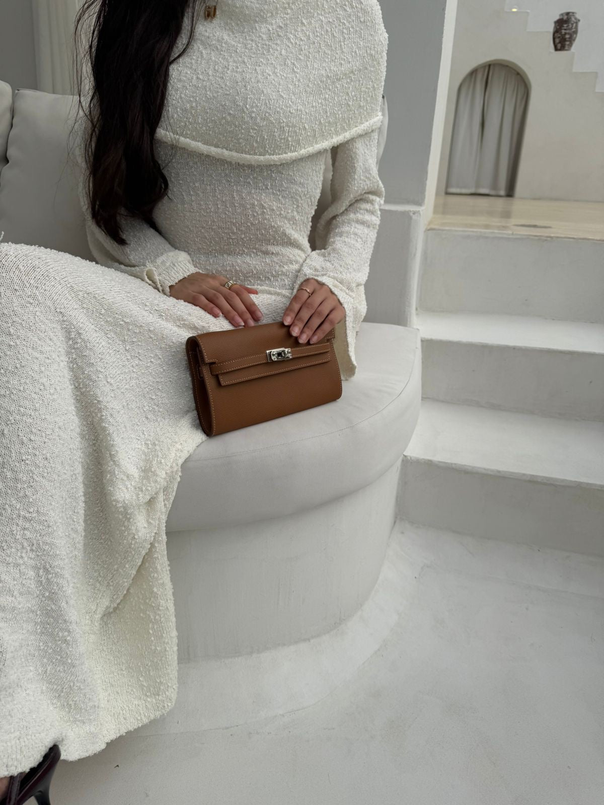 Woman in a white textured dress holding a brown handbag on a white chair.