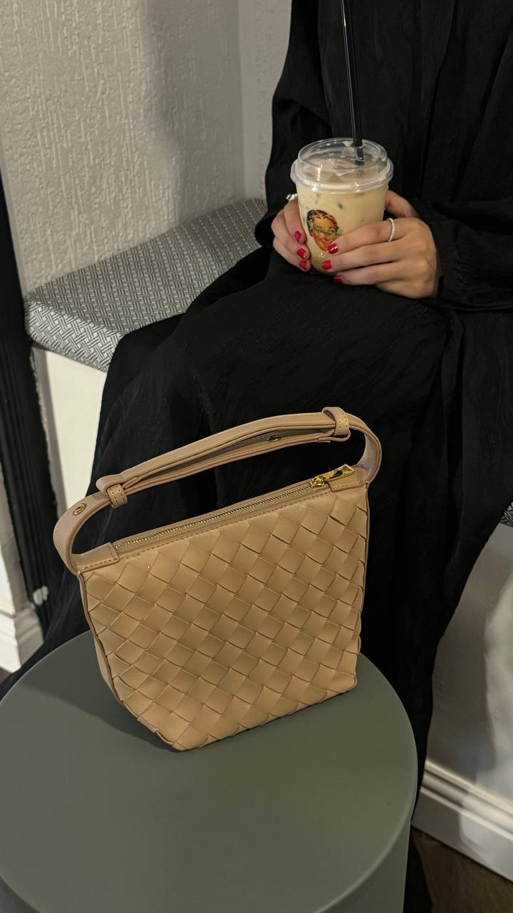 A beige handbag with a solid pattern placed on a table, with a person sitting in the background holding a cup.