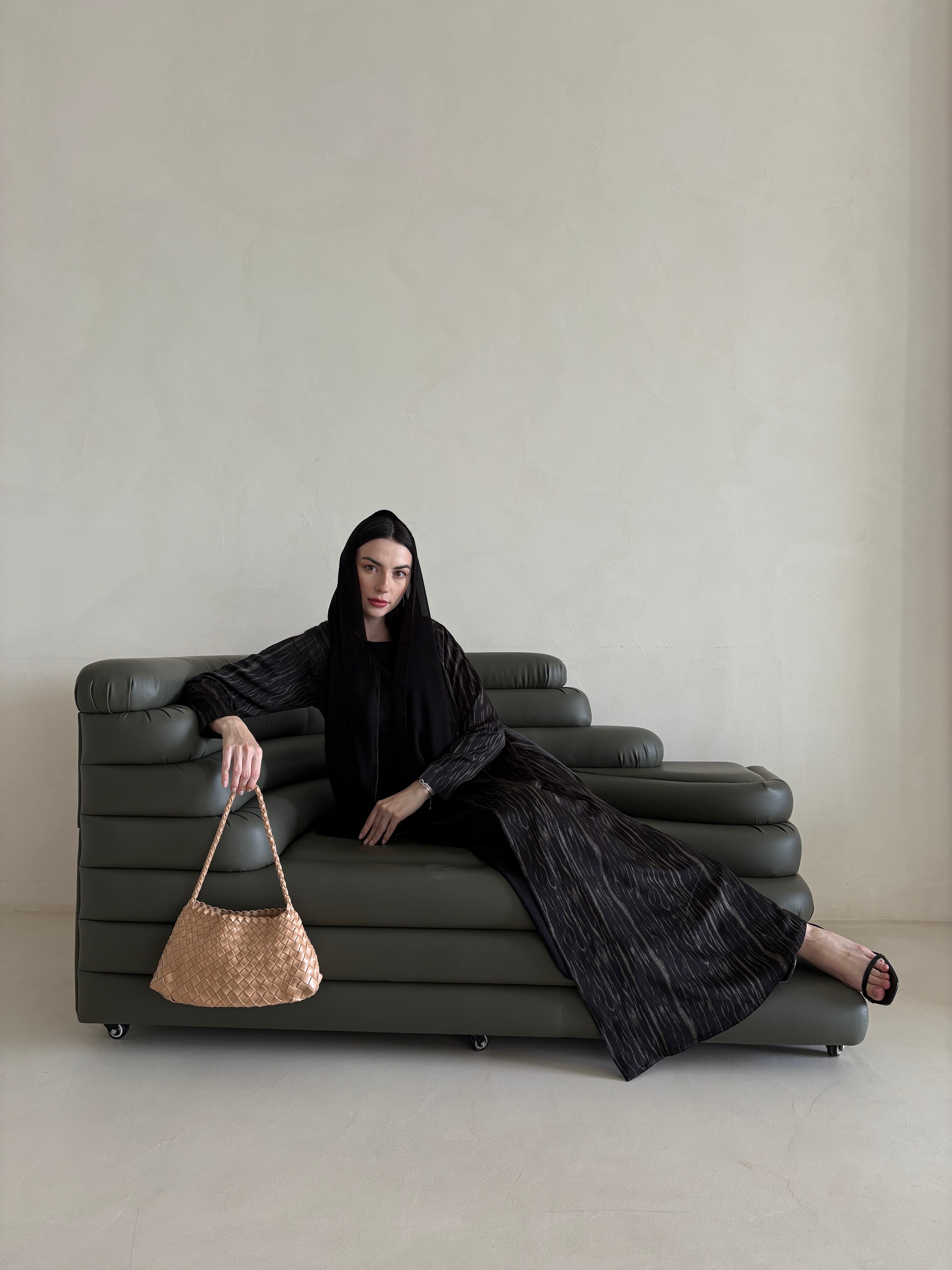 Woman in a black outfit sitting on a gray sofa holding a woven bag.