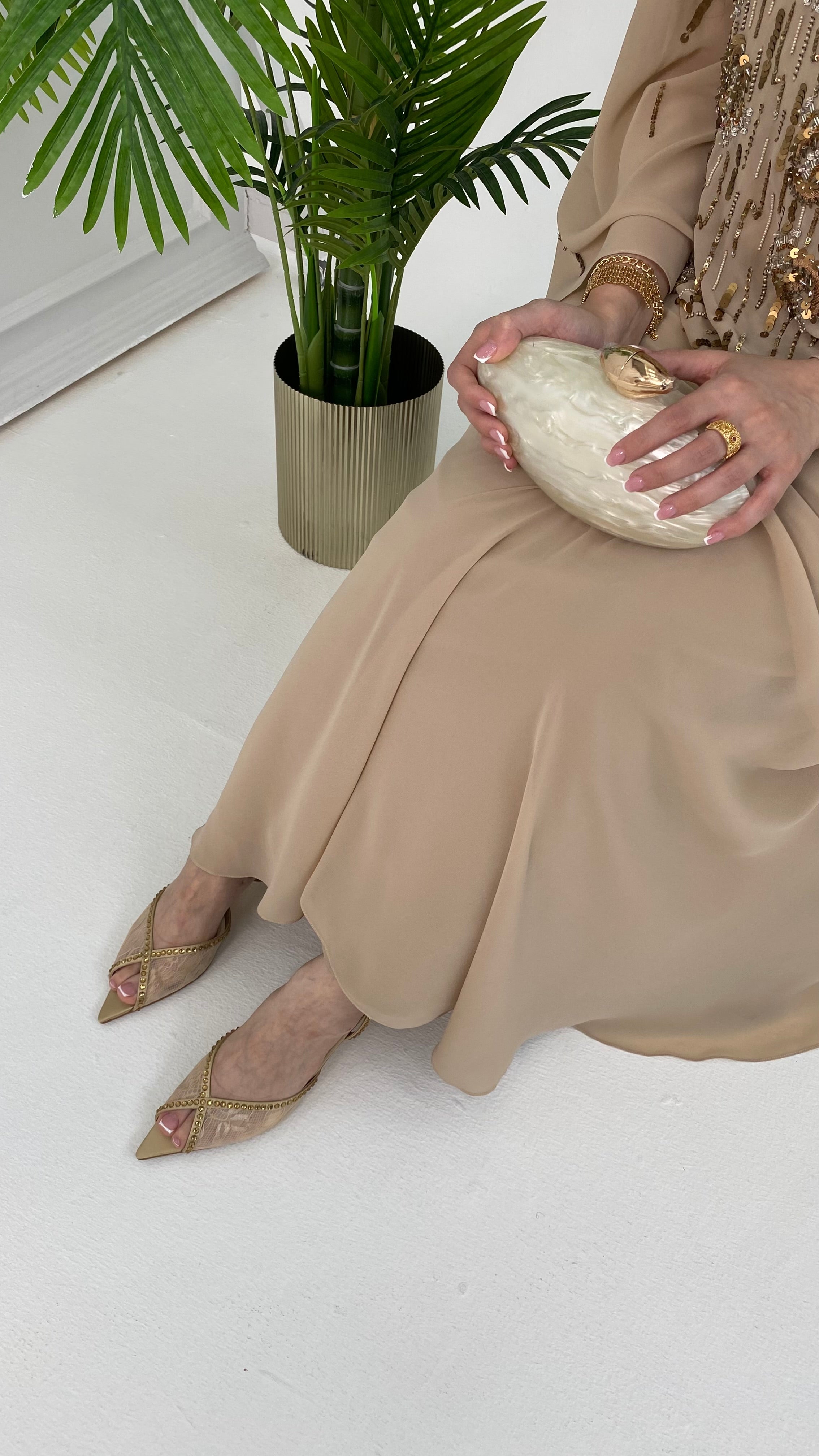Woman in a beige dress holding a white clutch with a plant in the background