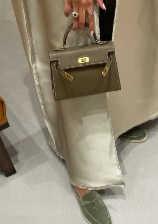 A small khaki handbag with a long strap being held by a person, showcased in a vertical orientation.