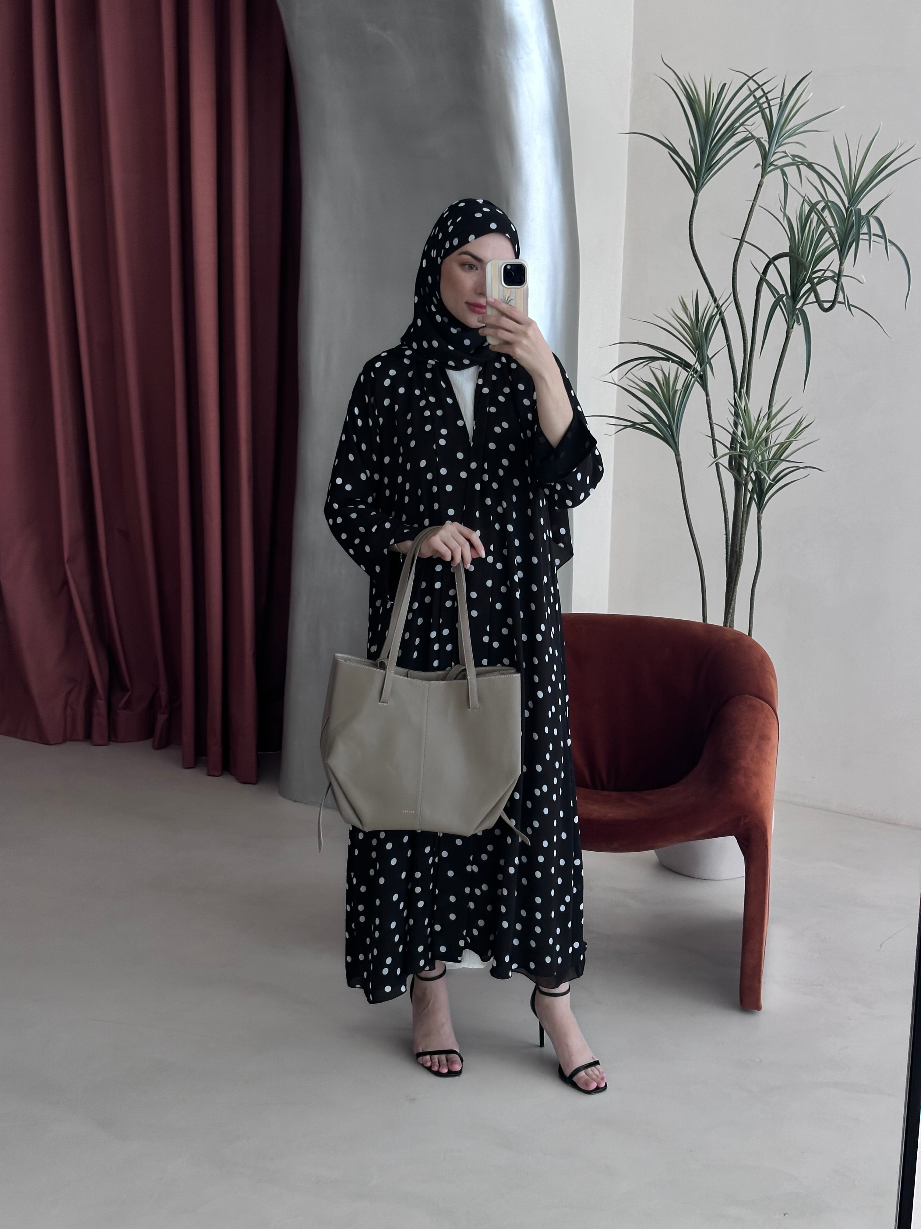 Woman in a polka dot dress holding a beige tote bag in a room with a plant and chair.