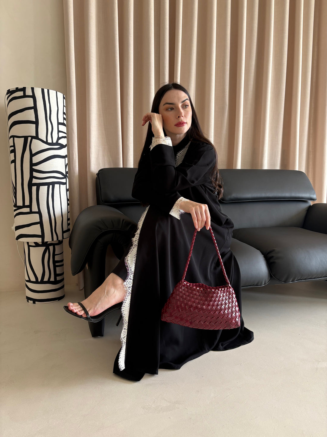 Woman in a black outfit holding a red handbag in a room with a black sofa and abstract sculpture.
