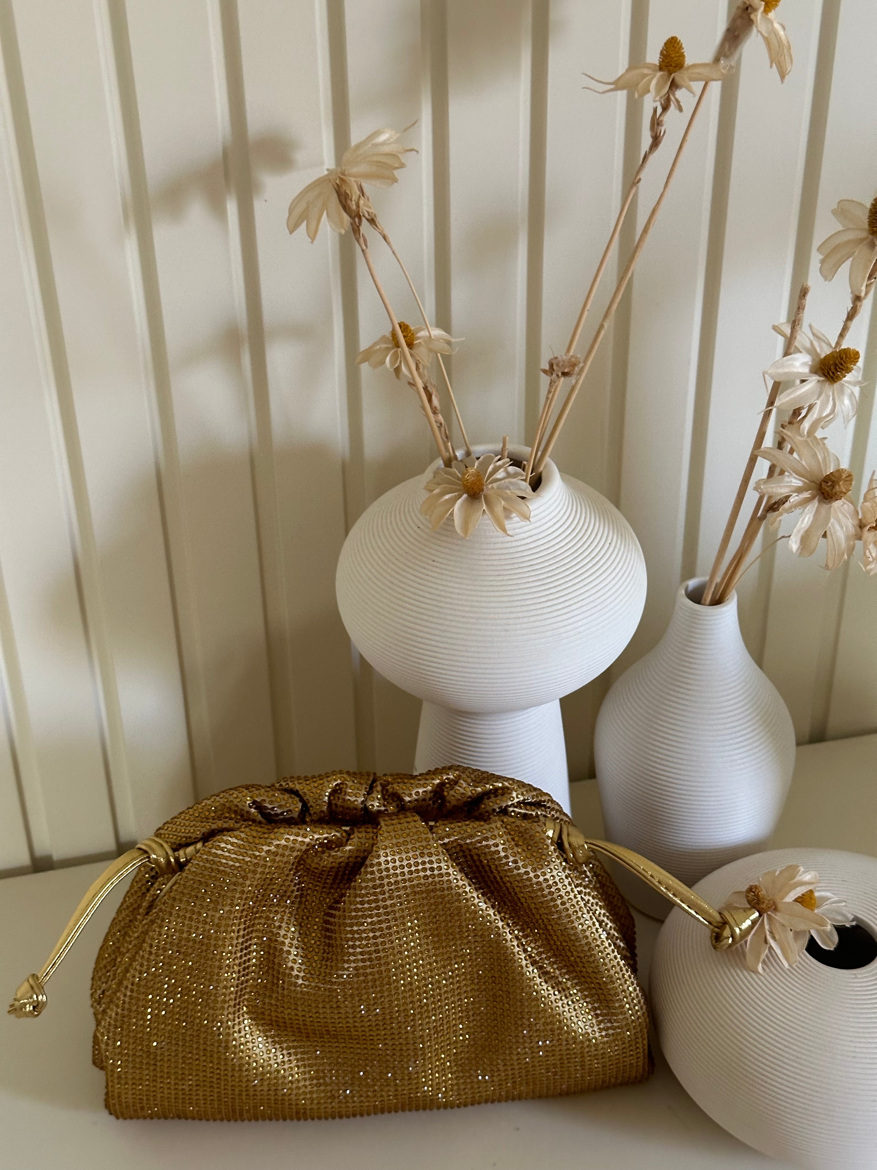 Gold purse next to white textured vases with dried flowers on a light wooden surface.