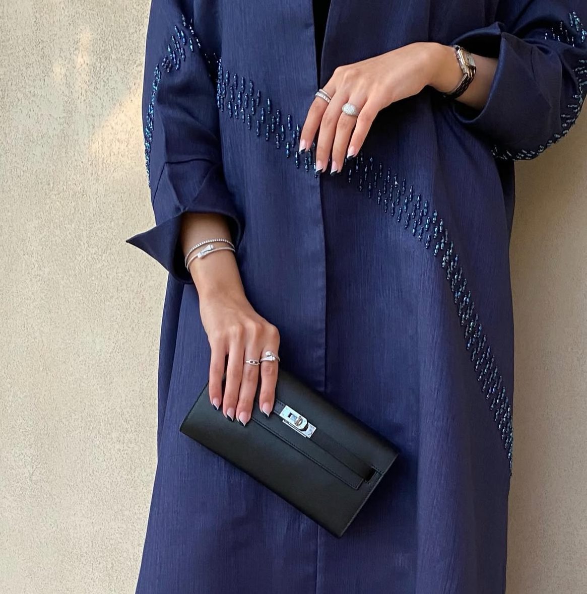 Person wearing a dark blue dress with intricate patterns, holding a black clutch against a neutral background.