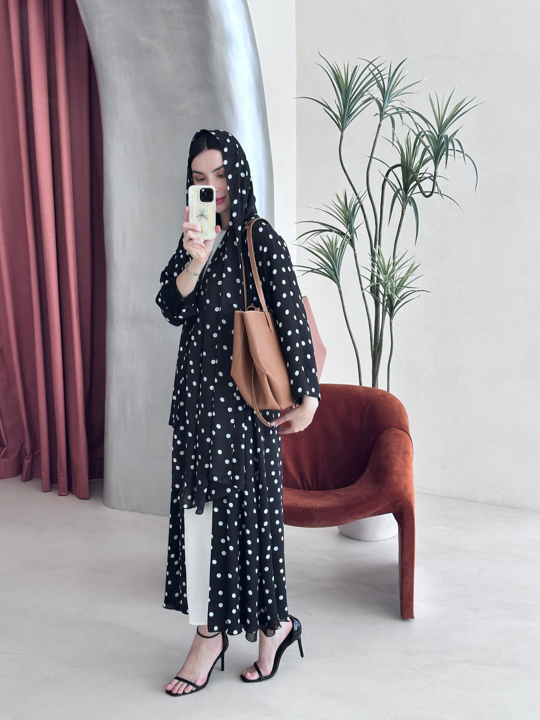 Person taking a mirror selfie wearing a black polka dot outfit with a brown bag in a room with pink curtains and a plant.