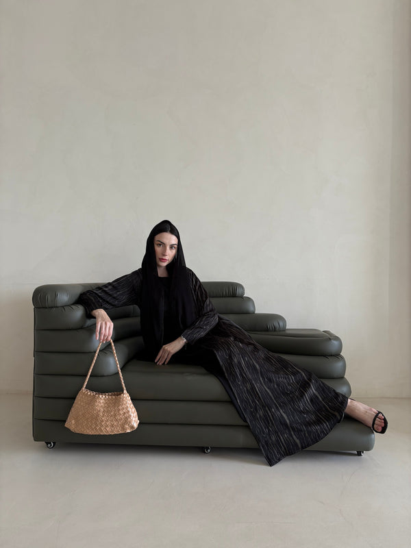 Woman in a black outfit sitting on a gray sofa holding a woven bag.