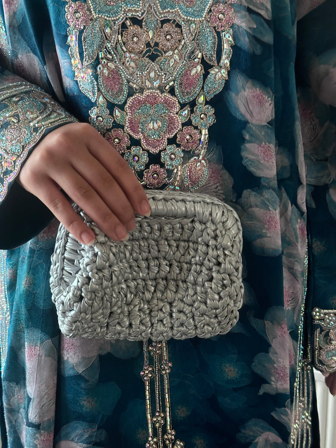 Hand holding a silver crochet handbag against a floral and embroidered background