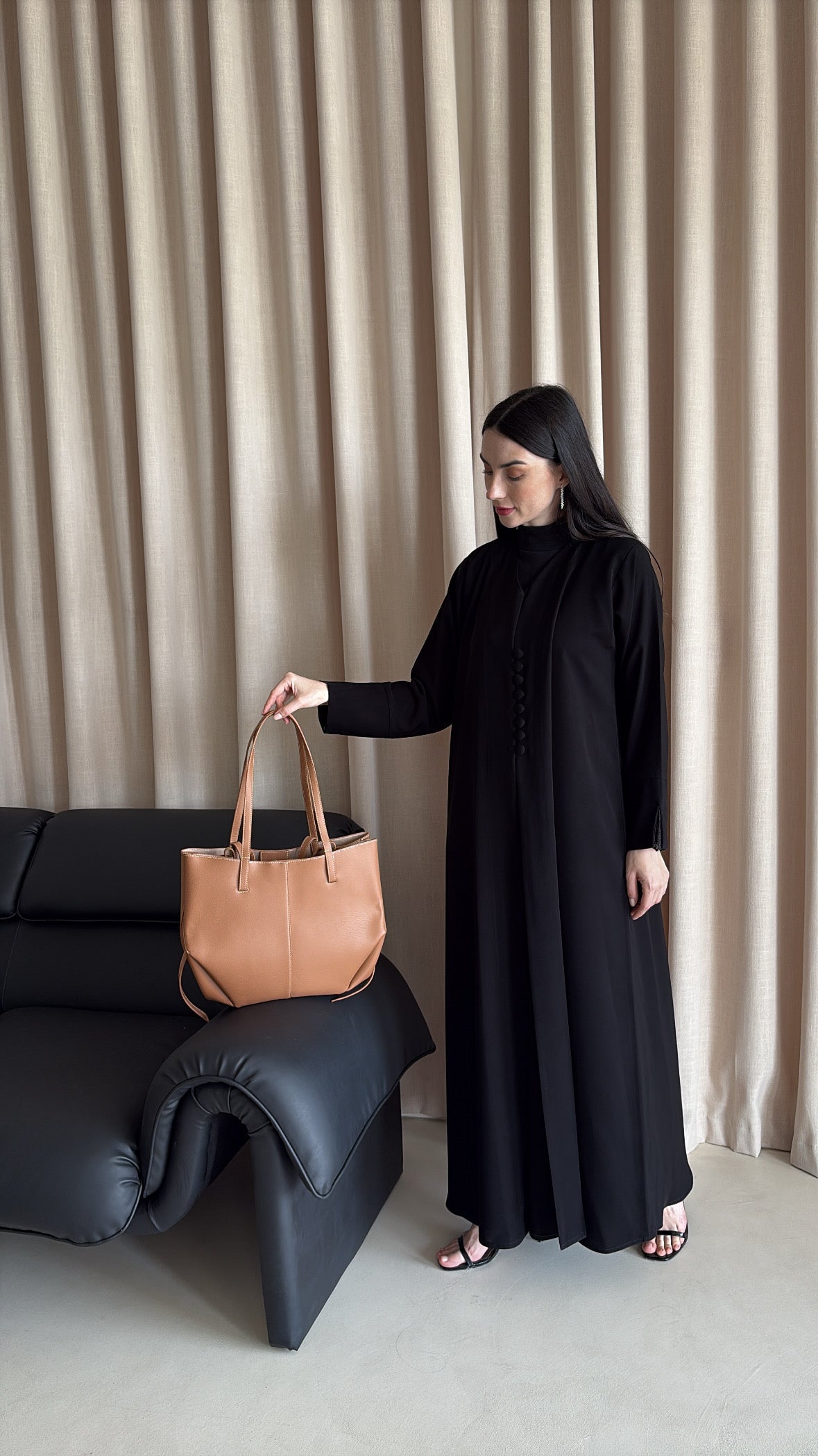Woman in a black dress holding a brown leather handbag against a beige curtain background