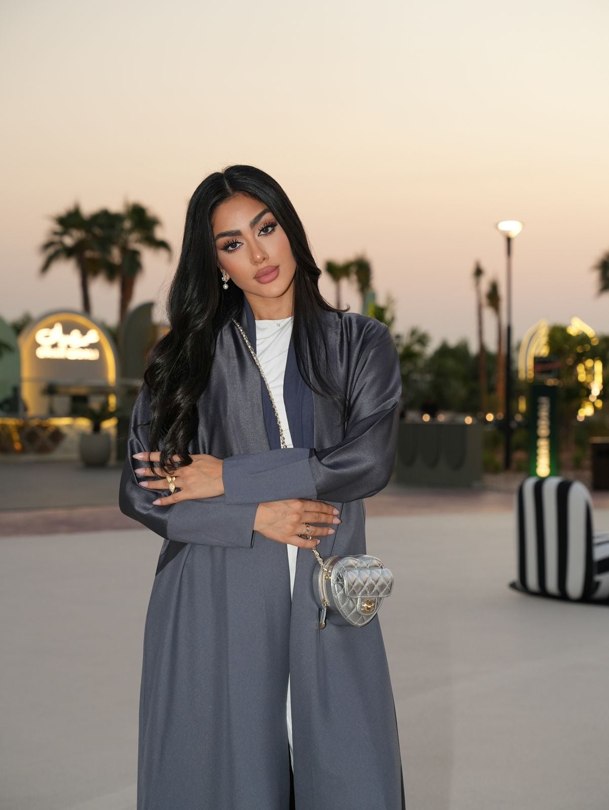 Woman in a gray outfit holding a handbag with palm trees and lights in the background