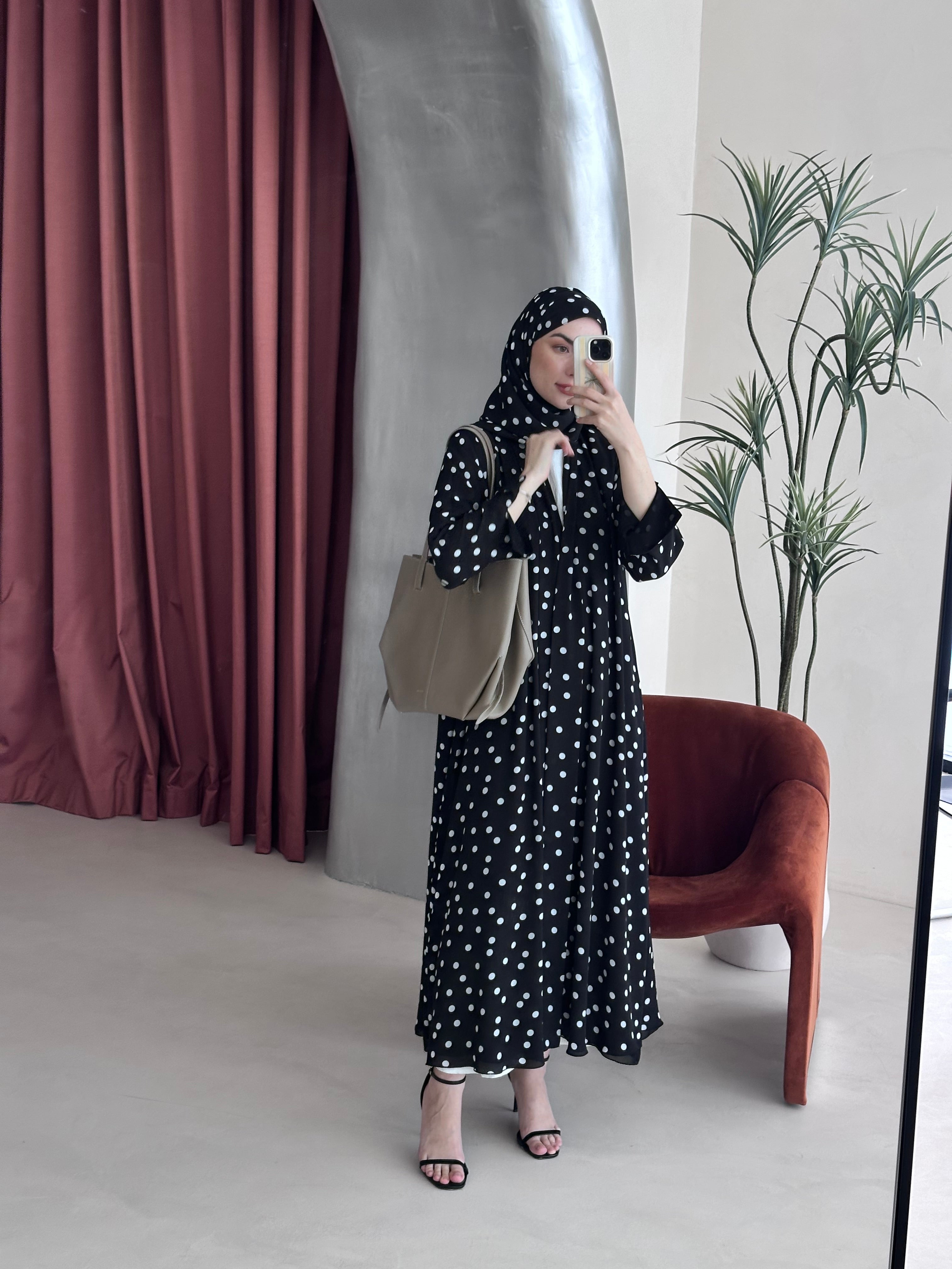 Woman in a black and white polka dot dress taking a mirror selfie in a room with a plant and chair.