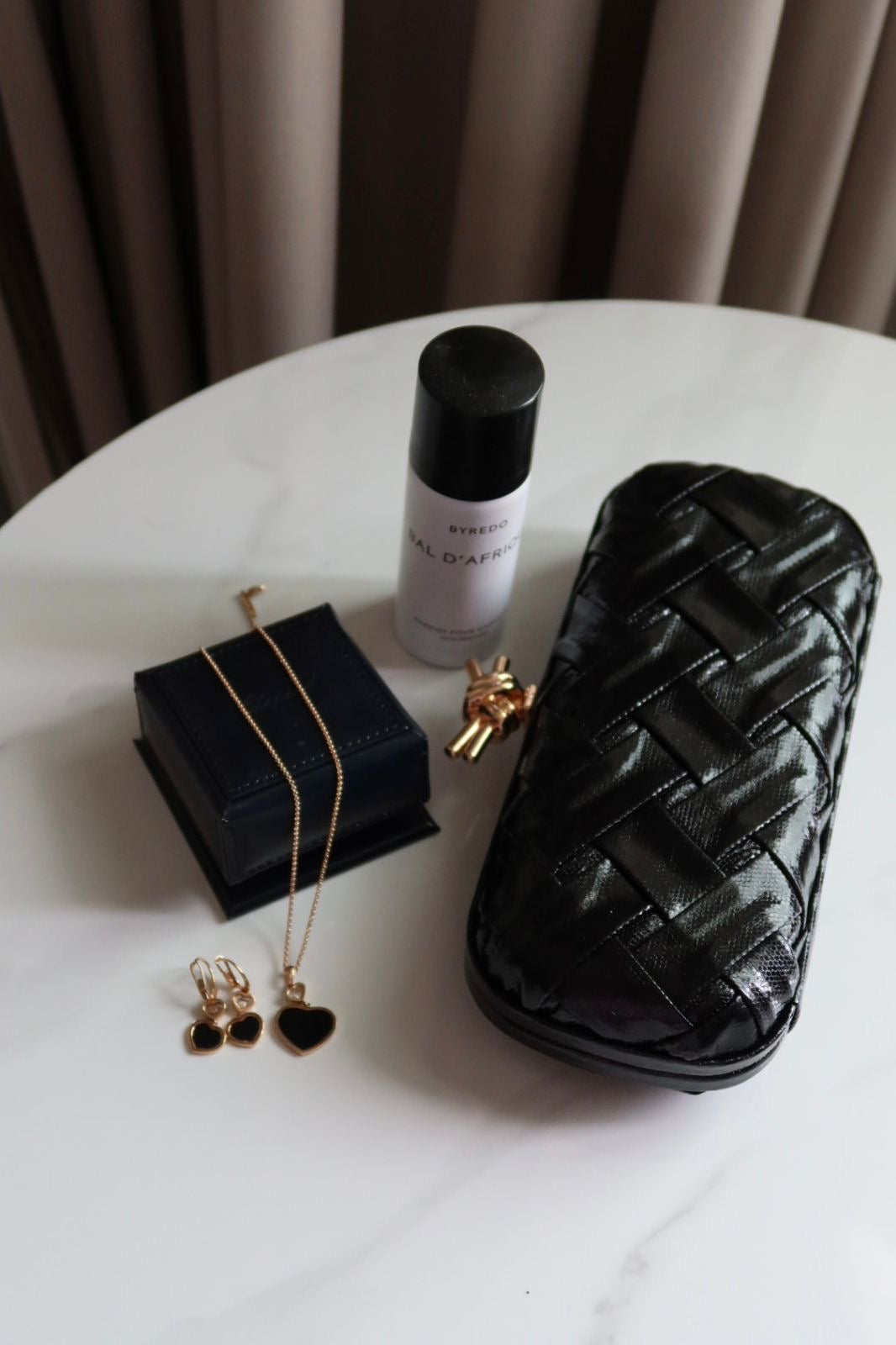 Black jewelry box, black and gold earrings, white bottle with black cap, and black woven pouch on a white surface.