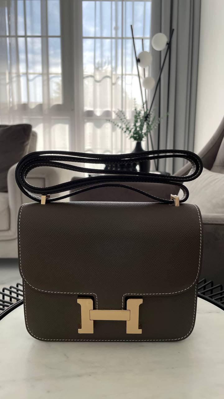 Brown handbag with gold H-buckle on a table in a room with large windows.