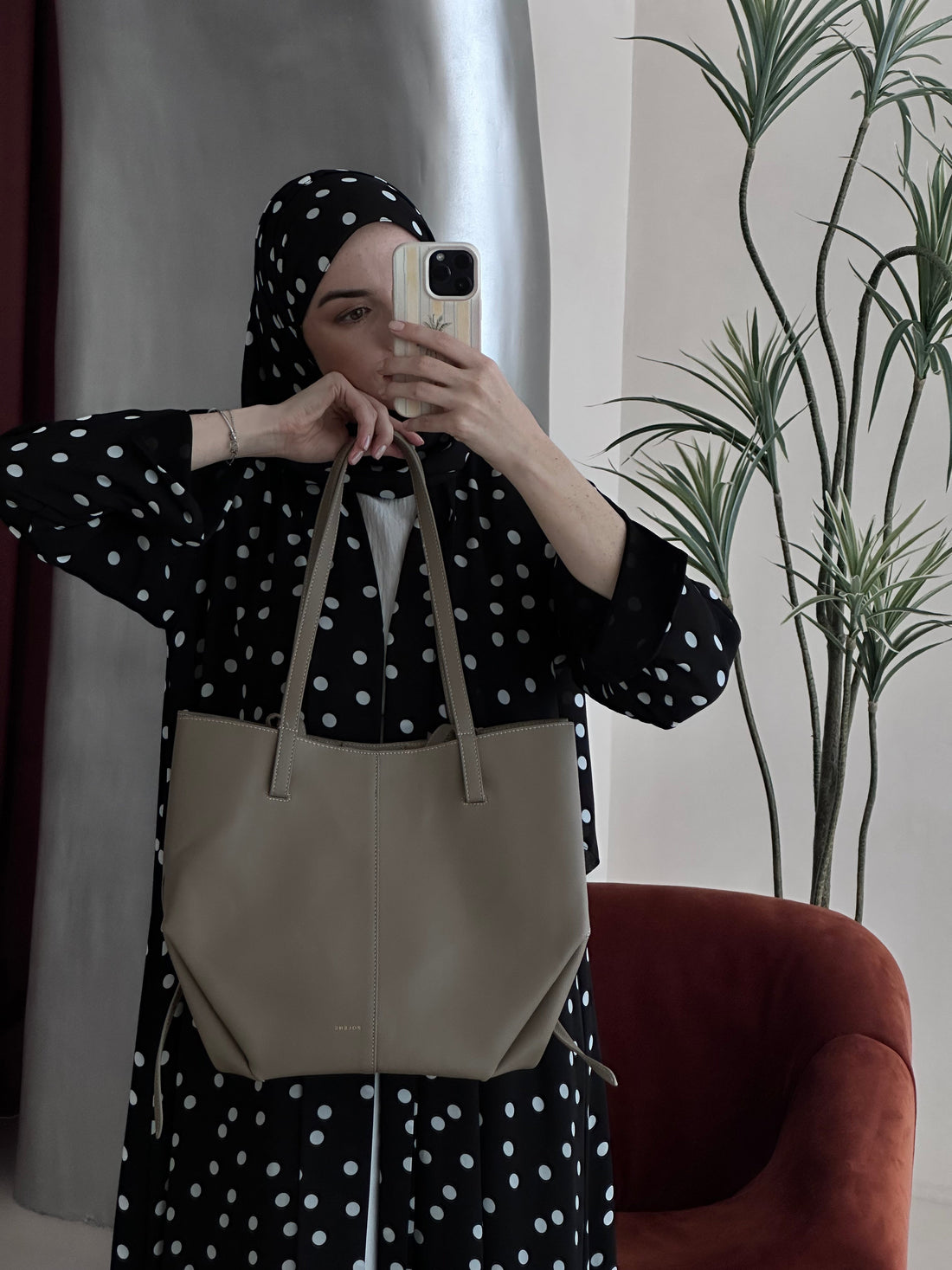 Person wearing a black polka dot outfit with a beige bag, taking a mirror selfie.